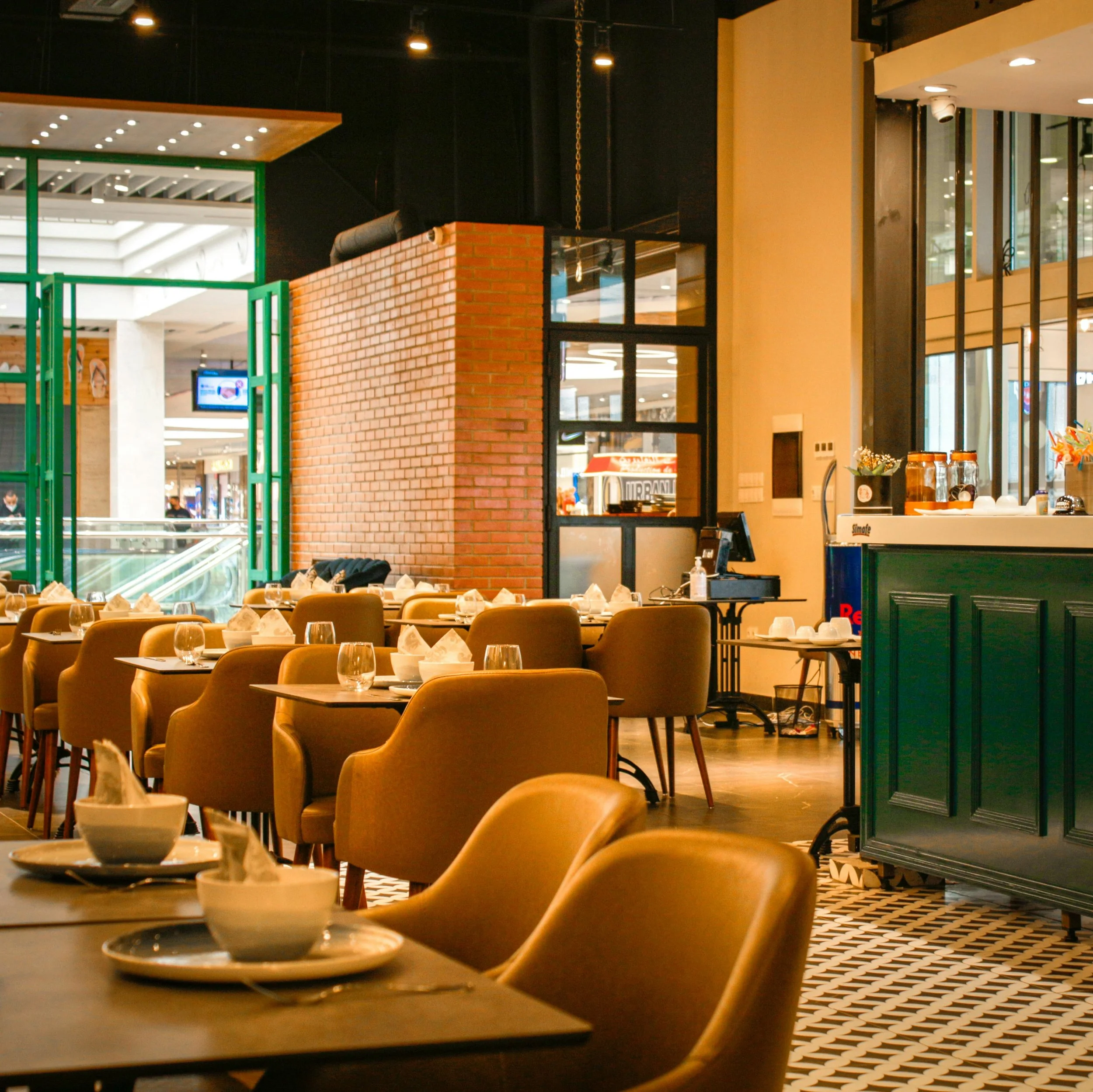 An outdoor restaurant patio overlooking a body of water with mountains in the background. The patio has tables set with plates, glasses, and utensils, and a partially open canopy with curtains.
