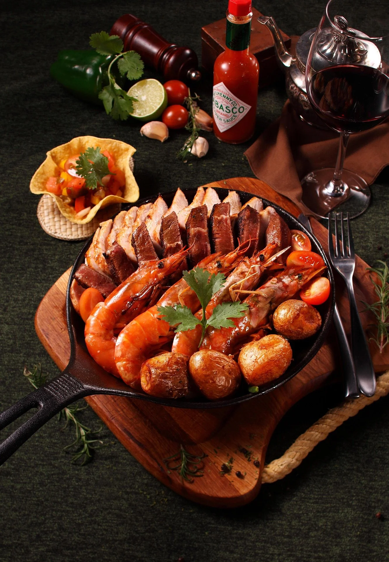 A cast iron skillet with cooked shrimp, steak slices, and roasted potatoes on a wooden board, surrounded by a glass of red wine, a lime, garlic, cherry tomatoes, hot sauce, and a side of pico de gallo.