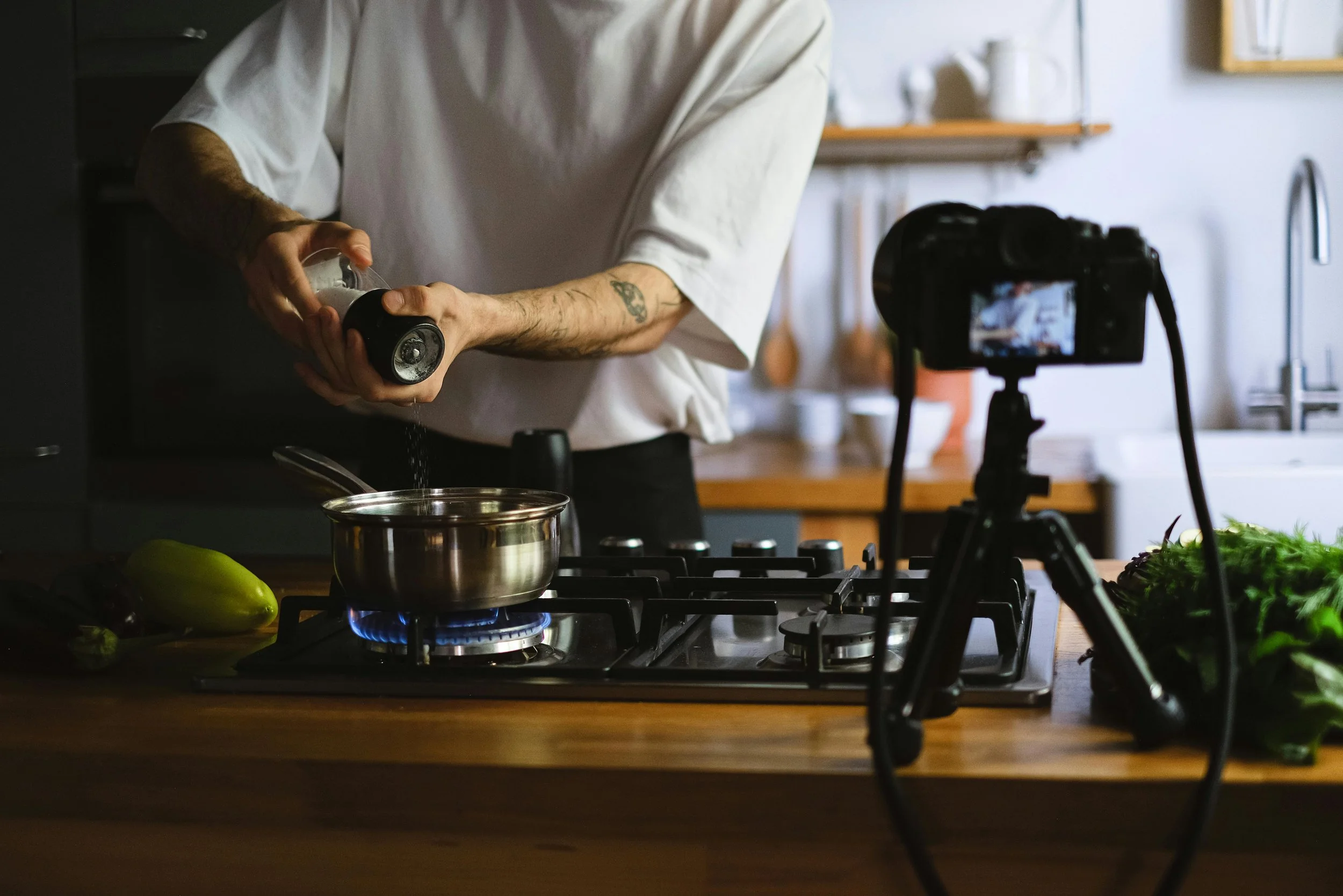 A person cooking on a stove while recording a cooking video with a camera on a tripod.
