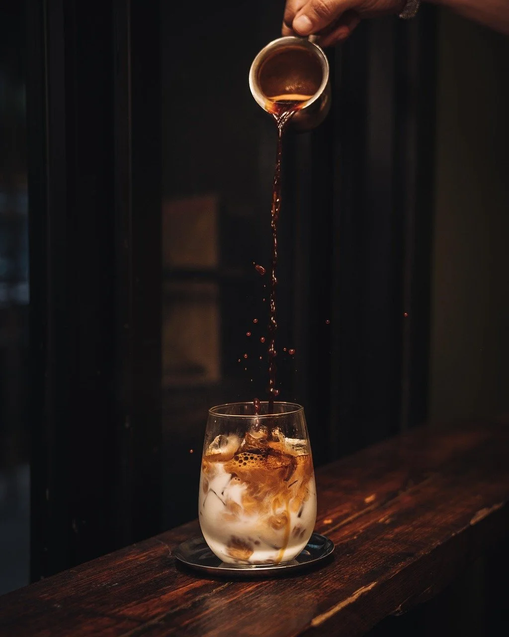 A hand pouring dark coffee or espresso into a glass of iced milk with ice cubes on a dark wooden surface.