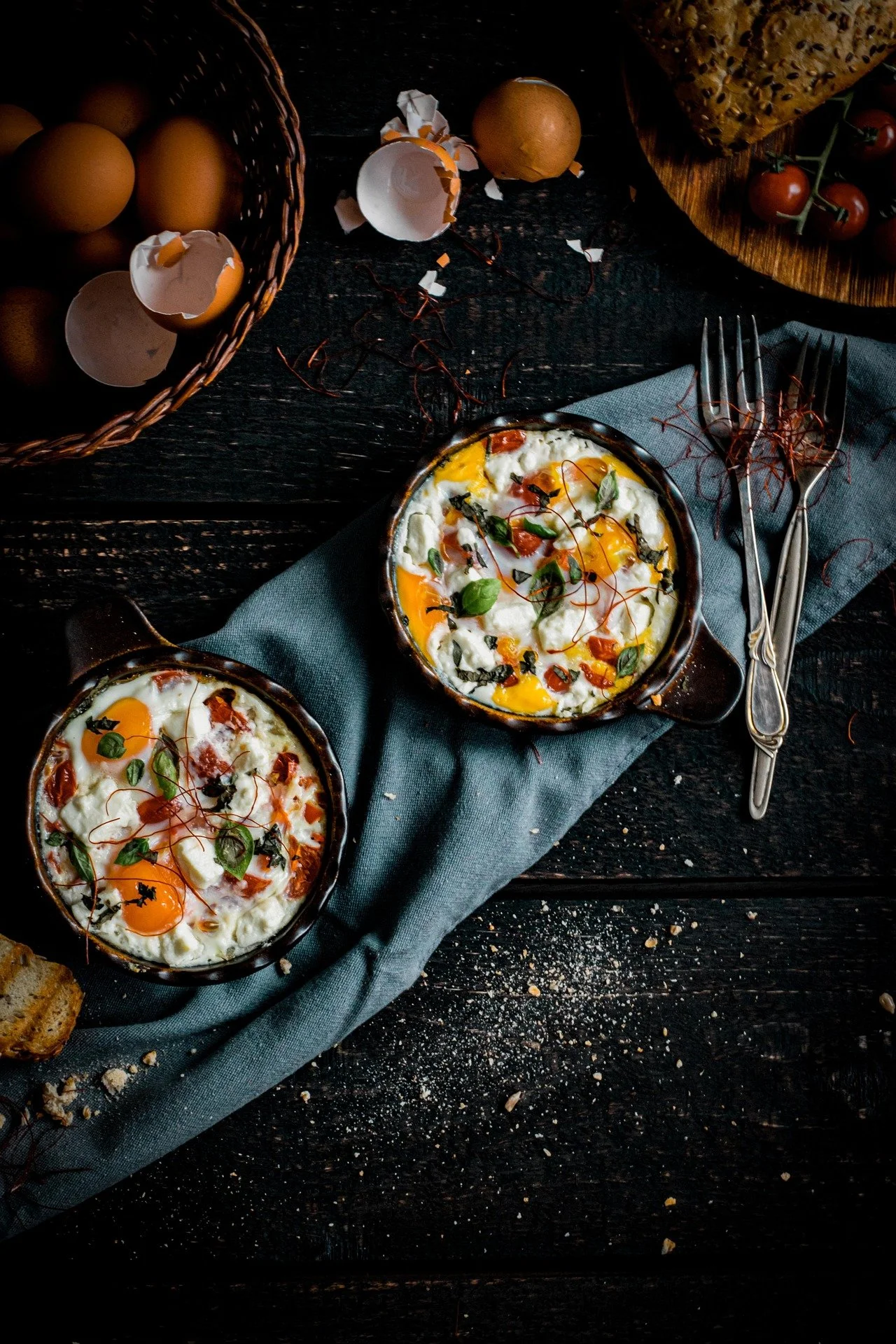 Two baked eggs with herbs and sauce in small black ramekins, laid on a dark wooden table with a blue cloth, surrounded by eggs, broken eggshells, cherry tomatoes, a loaf of bread, and forks.