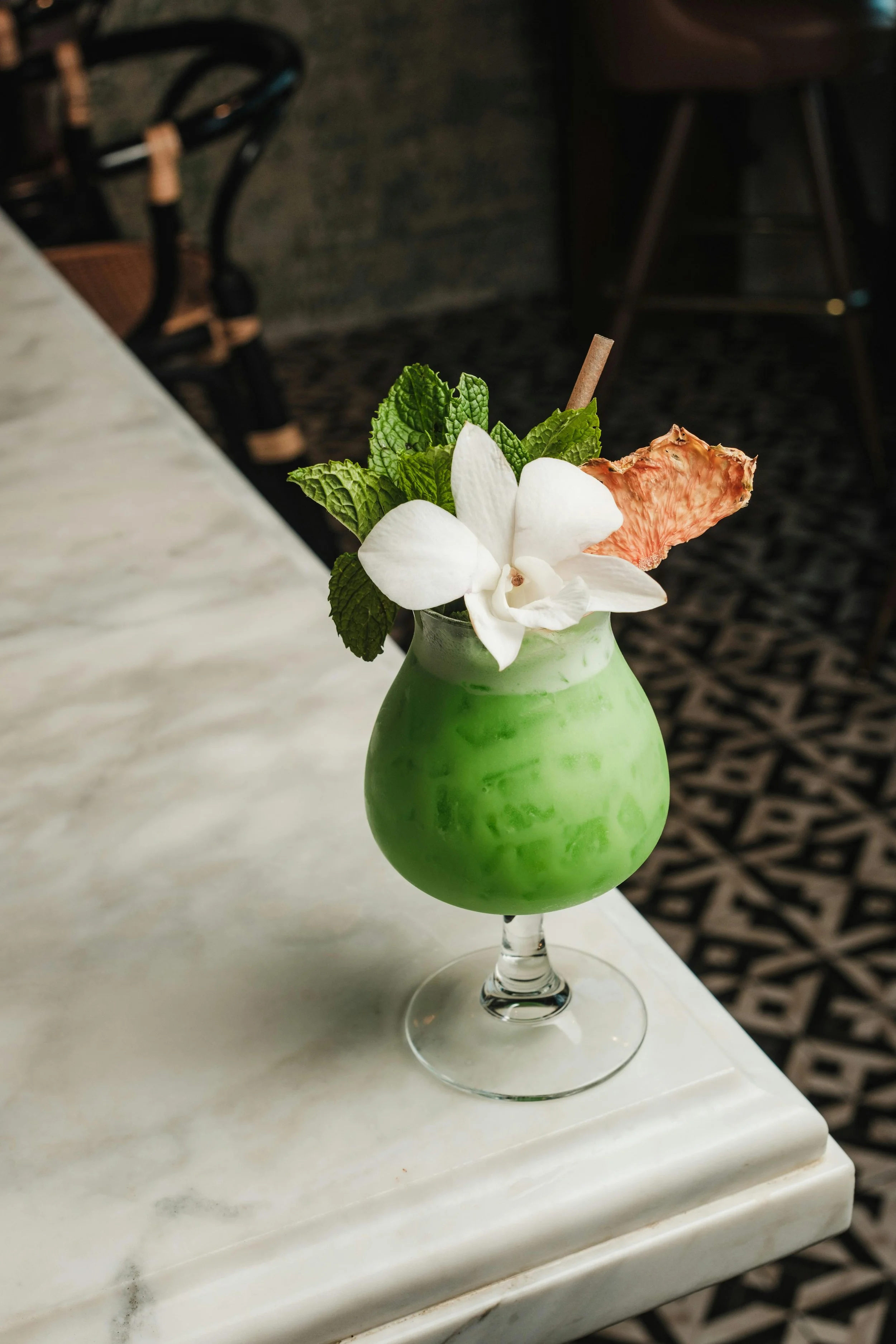 A green coconut drink topped with white flower, mint leaves, dried fruit, and a straw, served in a stemmed glass on a white marble table.