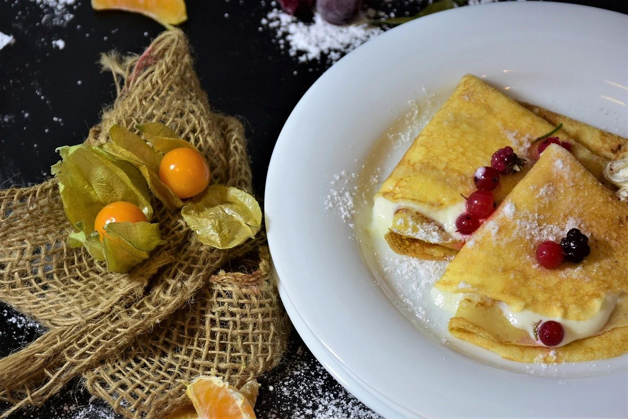 A white plate with three crepes filled with creamy filling, topped with powdered sugar and red and black berries. Next to the plate, a decorative bunch of Physalis fruits with orange berries and papery husks is placed on a black surface.