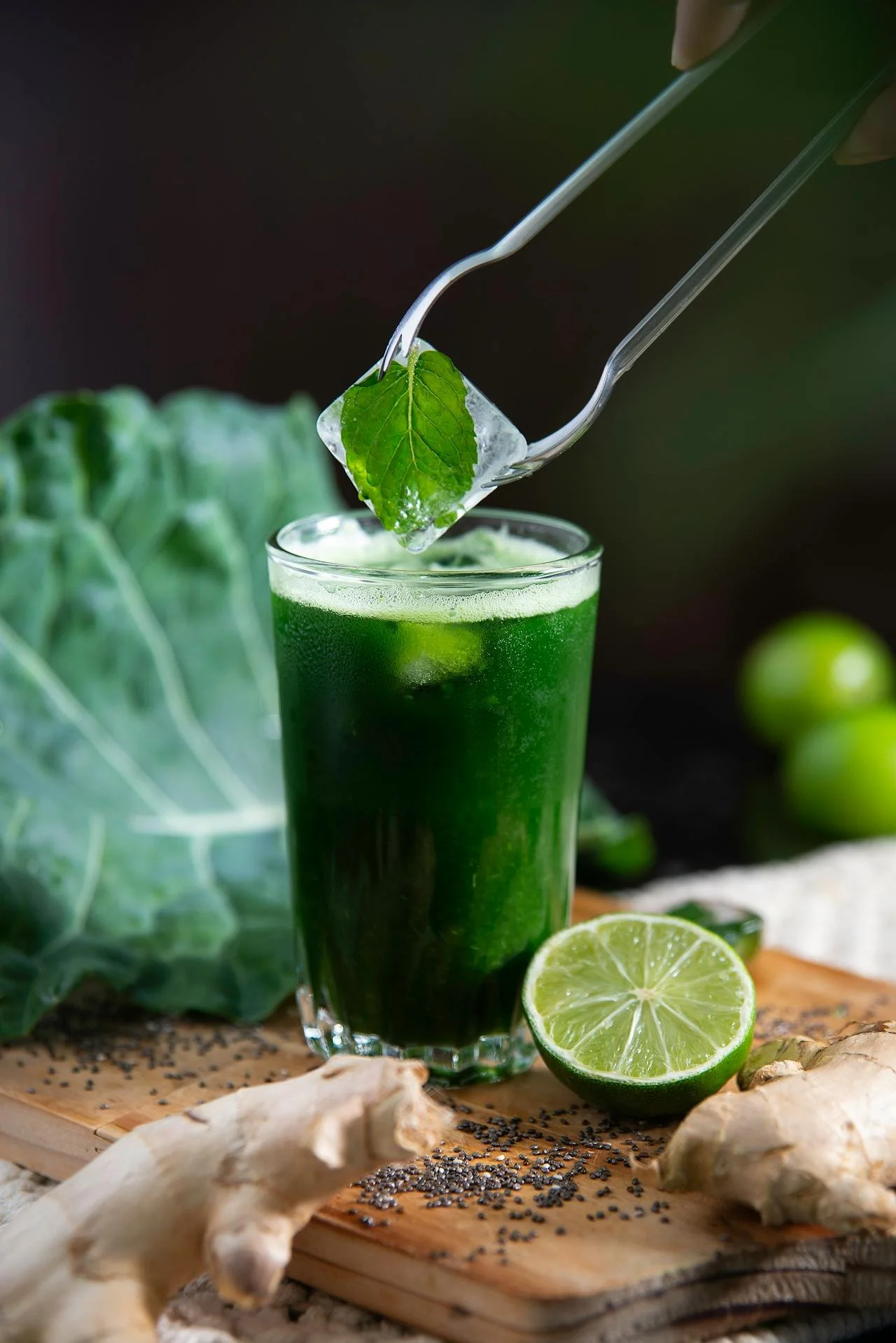 A tall glass of green juice with lime, ginger, and leafy greens, with a metal tong holding an ice cube and a leaf above the glass, surrounded by ingredients like ginger, lime, and black seeds on a wooden cutting board.