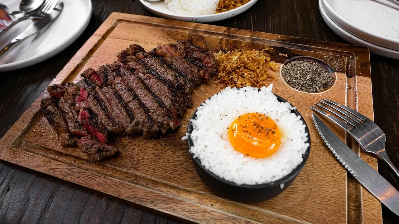 A plate with grilled steak, a bowl of white rice topped with an egg yolk, shredded meat or vegetables, and two small containers of condiments on a wooden serving board.