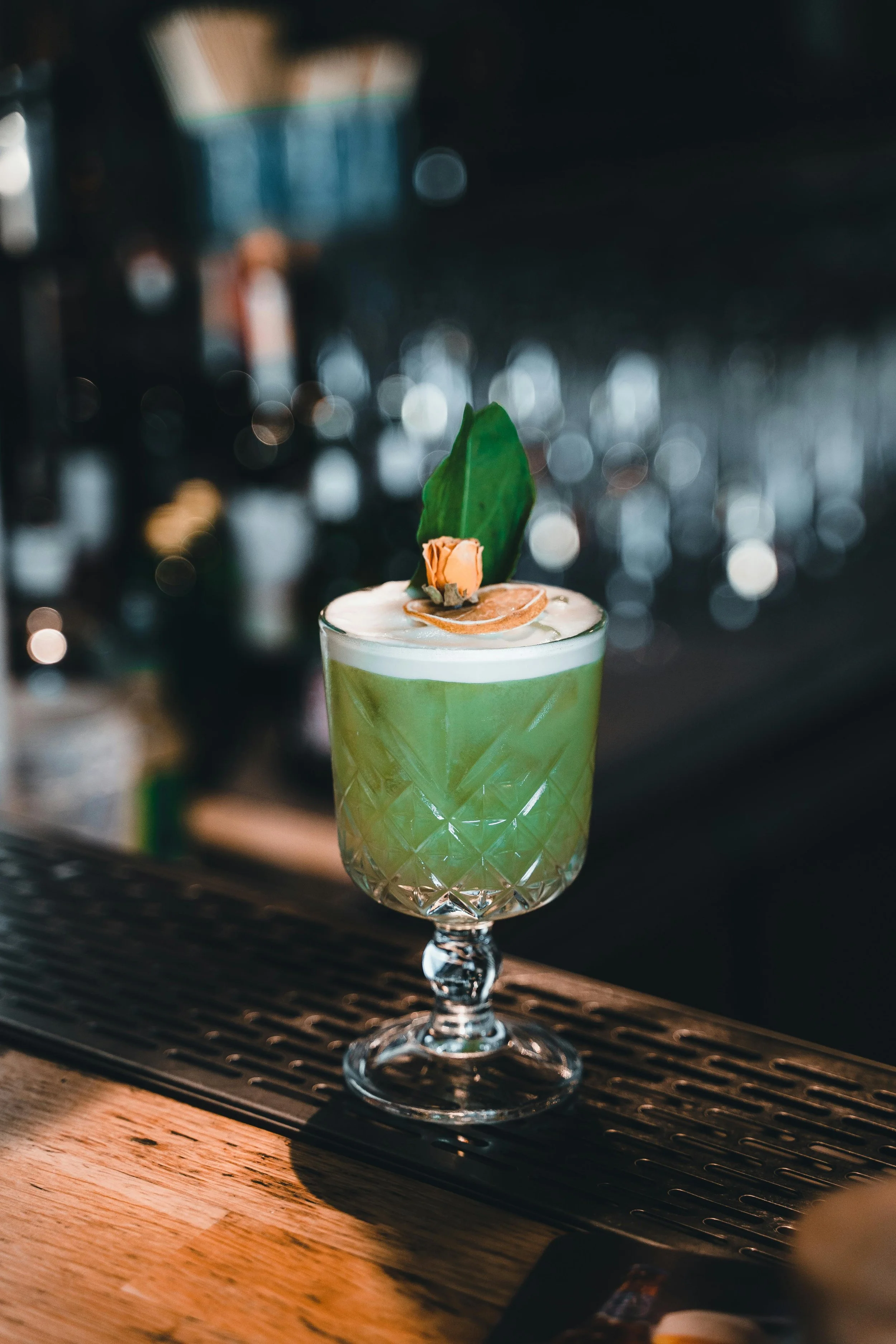 A green cocktail in a textured glass garnished with a leaf and a dried orange slice, sitting on a wooden bar counter.