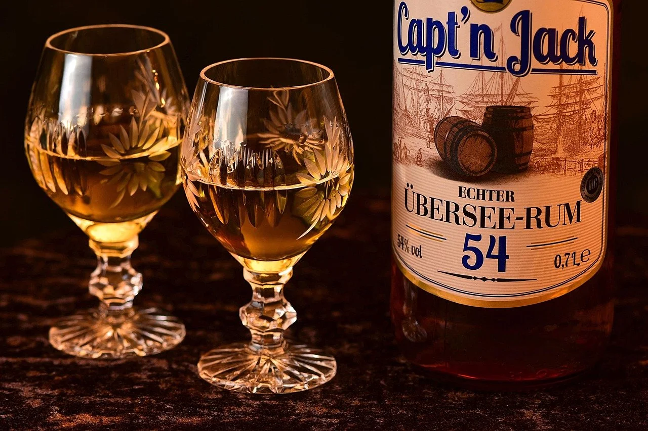 Two glasses of amber-colored rum on a dark surface next to a bottle labeled 'Captain Jack Übersee-Rum' with an illustration of barrels and ships.