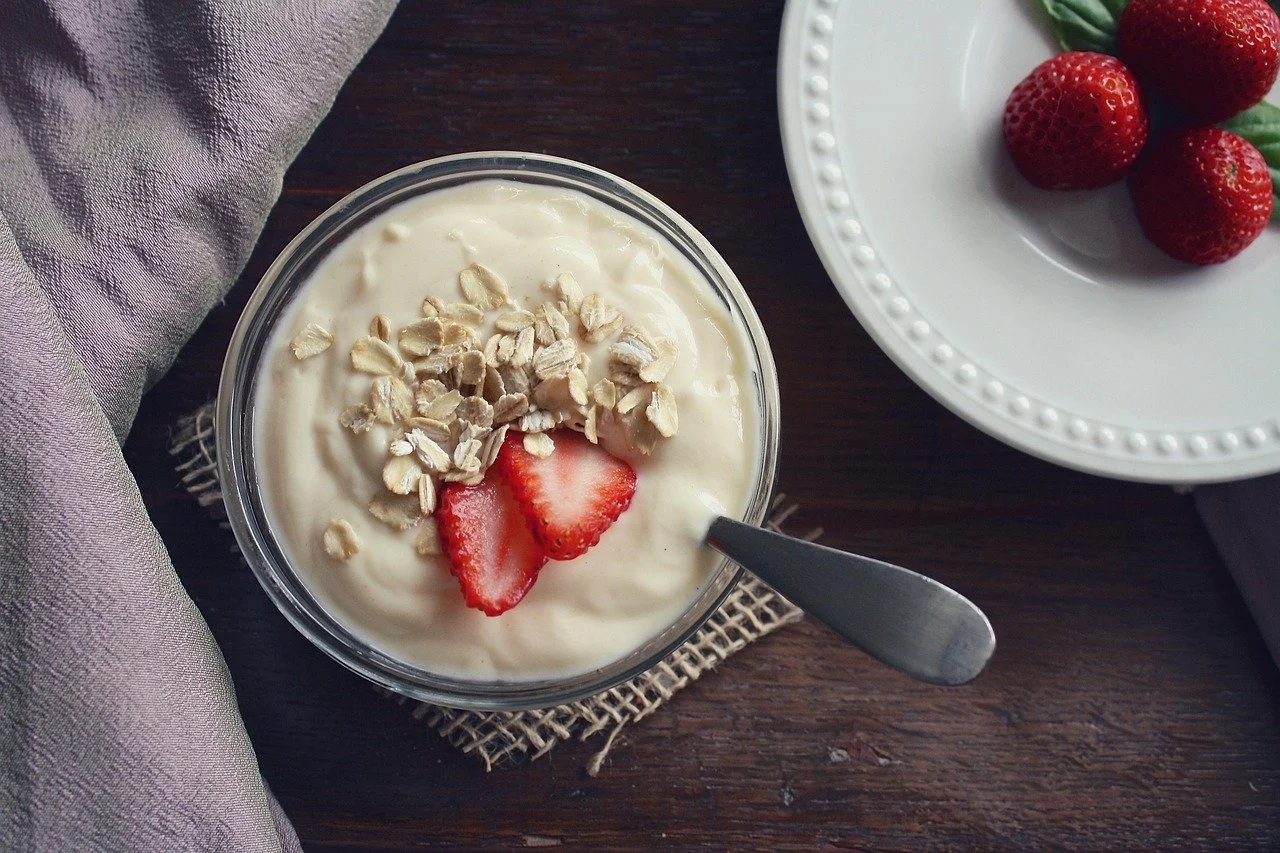 A glass bowl of yogurt topped with sliced strawberries and chopped oats on a dark wooden table. A white plate with strawberries is partially visible on the right side of the image.