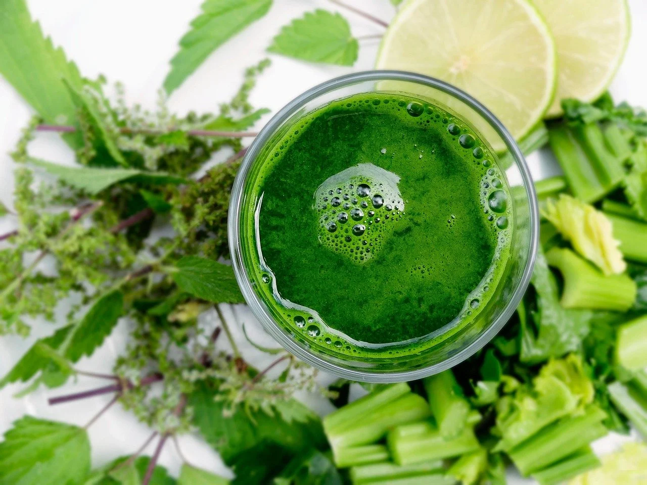 A glass of green smoothie surrounded by fresh herbs, lime slices, and chopped celery.
