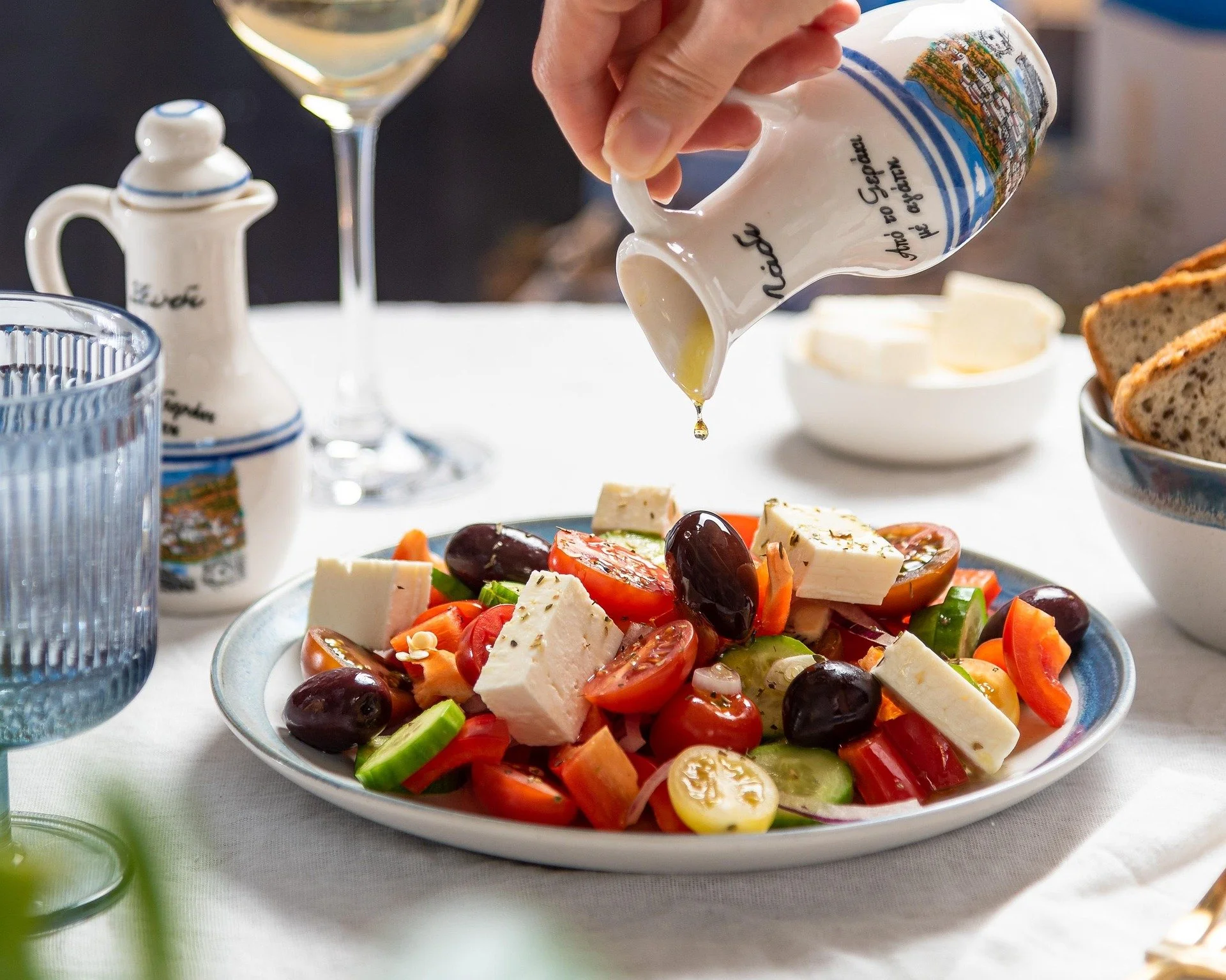 A person pouring olive oil onto a Greek salad with tomatoes, cucumbers, olives, and feta cheese, on a white table with wine, bread, and condiments in the background.