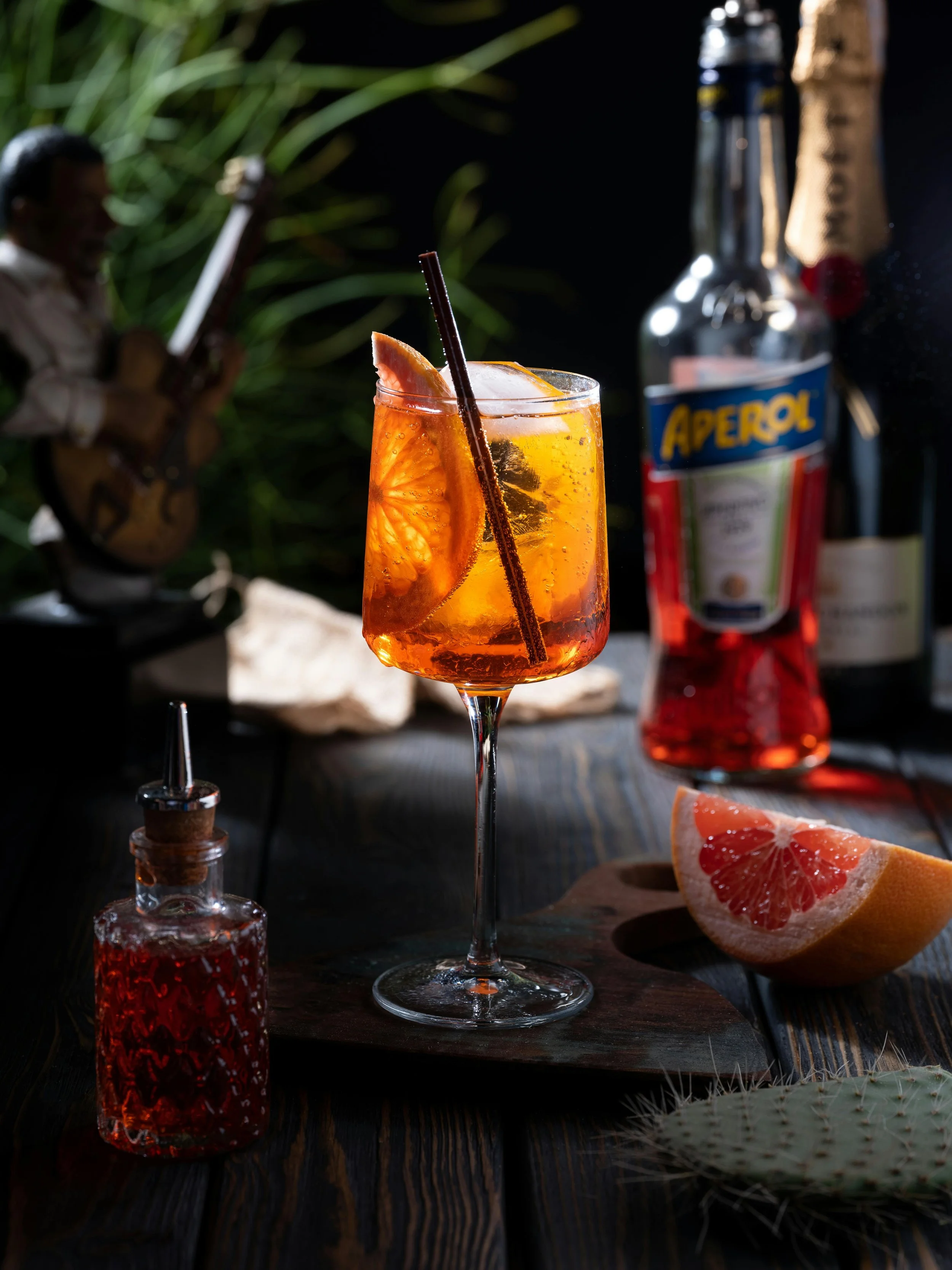 A cocktail garnished with a grapefruit slice and a straw on a dark wooden surface, with bottles of Aperol and liquor, a grapefruit wedge, and a cactus in the background.
