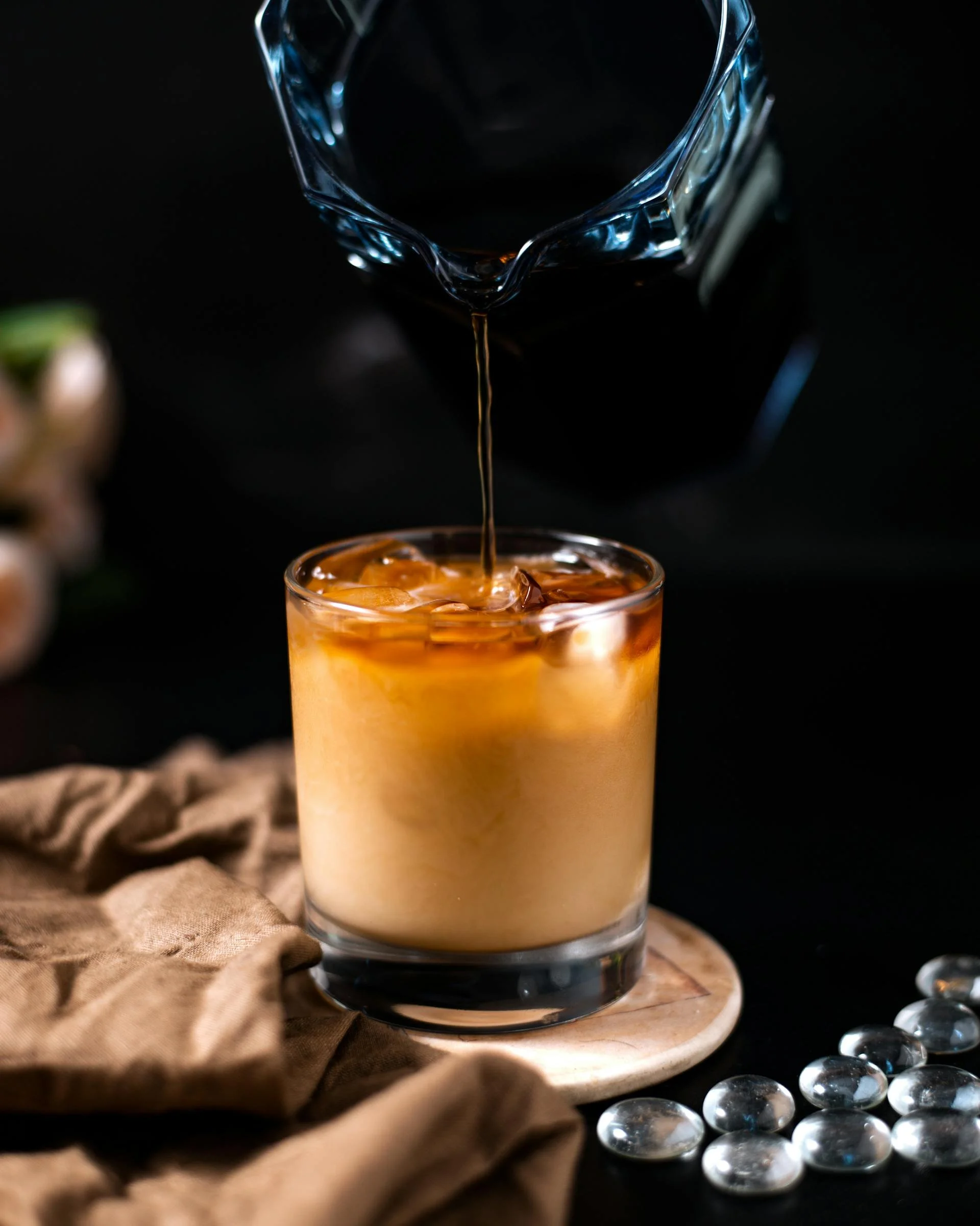 A glass of iced coffee with milk on a wooden coaster, with dark coffee being poured into it from a glass carafe, on a black background with a brown cloth and decorative glass stones nearby.