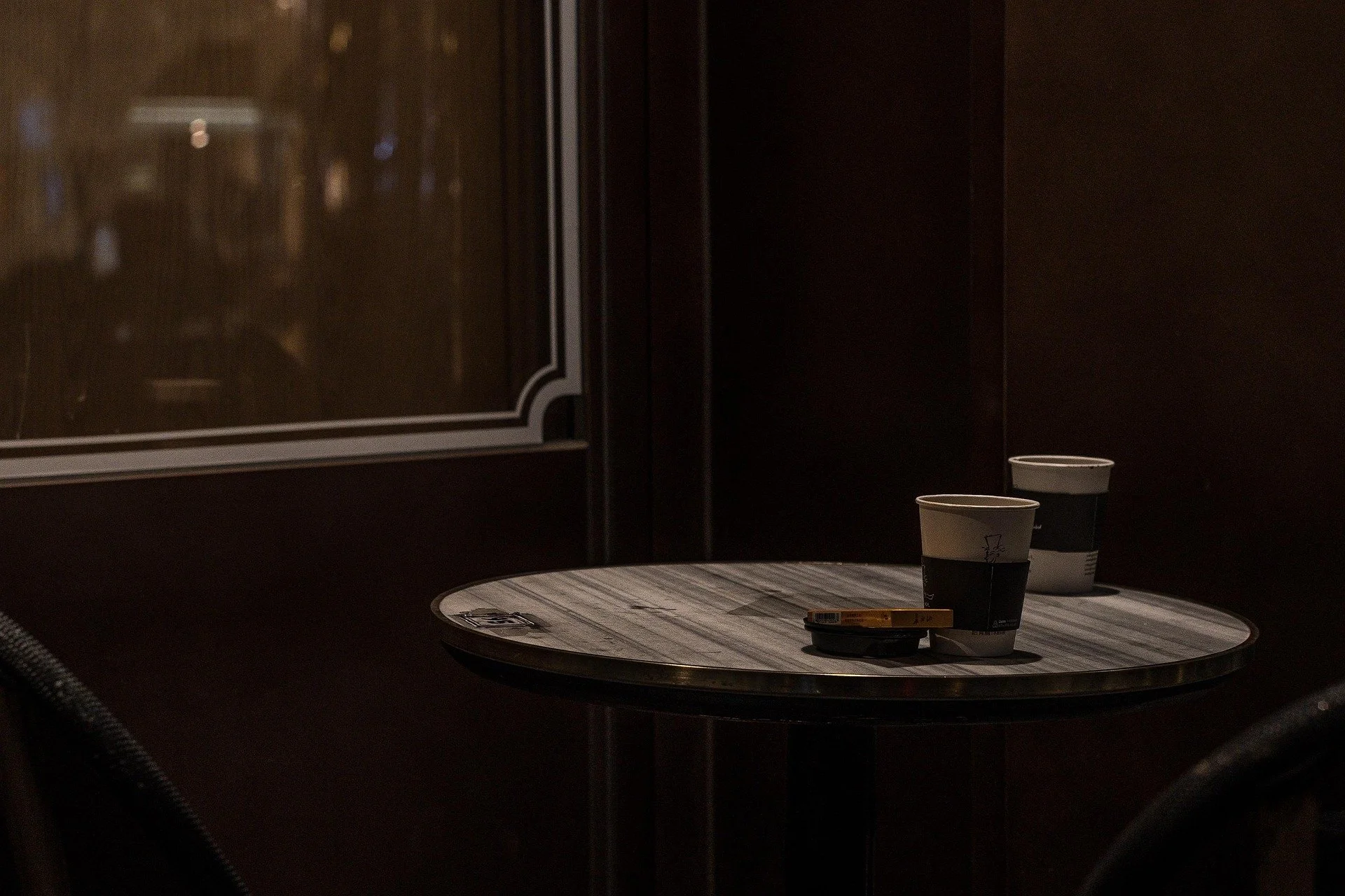 A small round table with two coffee cups and a pack of cigarettes in a dimly lit cafe or restaurant.