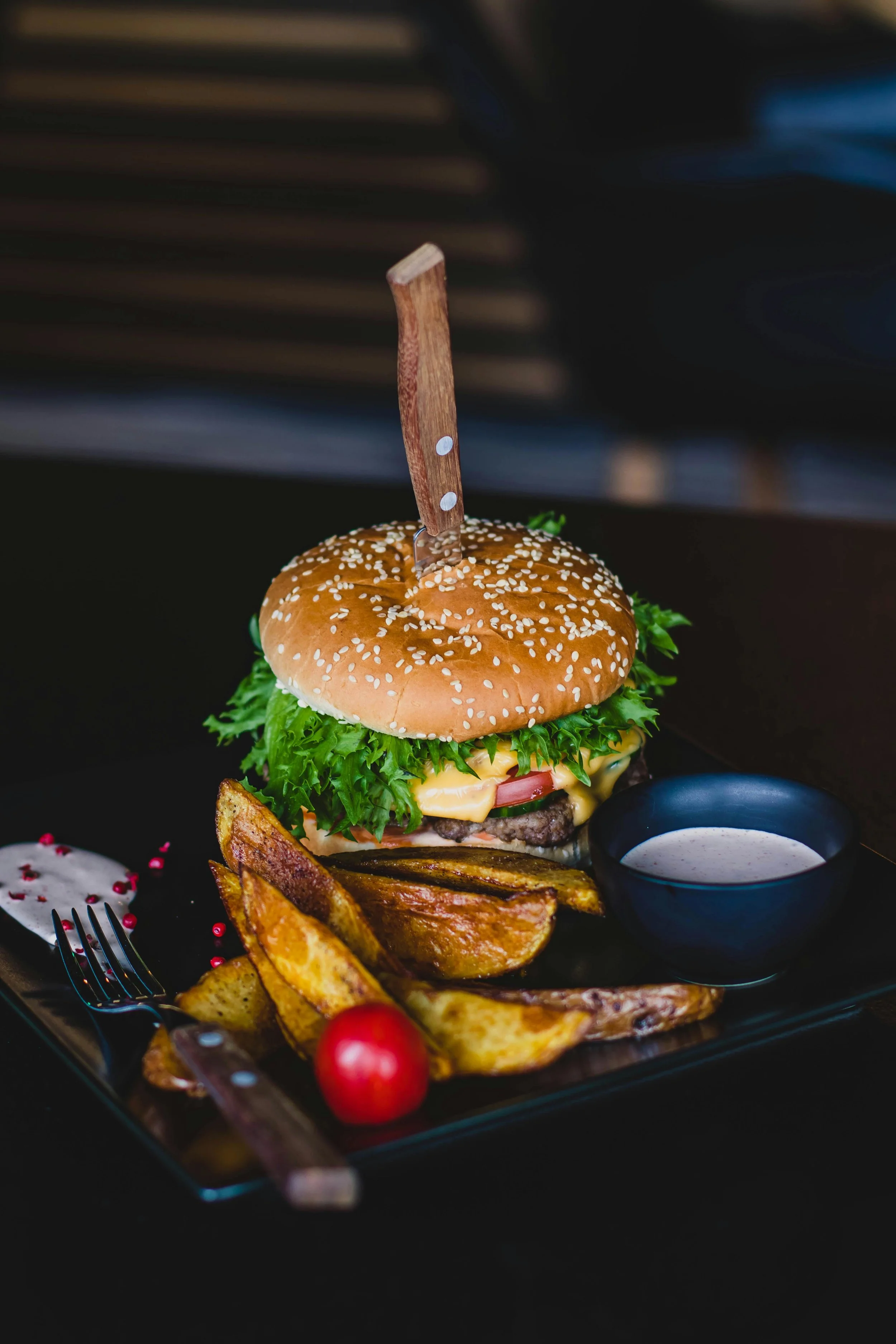 A cheeseburger with lettuce, tomato, cheese, and a sesame seed bun, served with potato wedges, a cherry tomato, and a small bowl of dipping sauce on a black plate with decorative pink peppercorns.