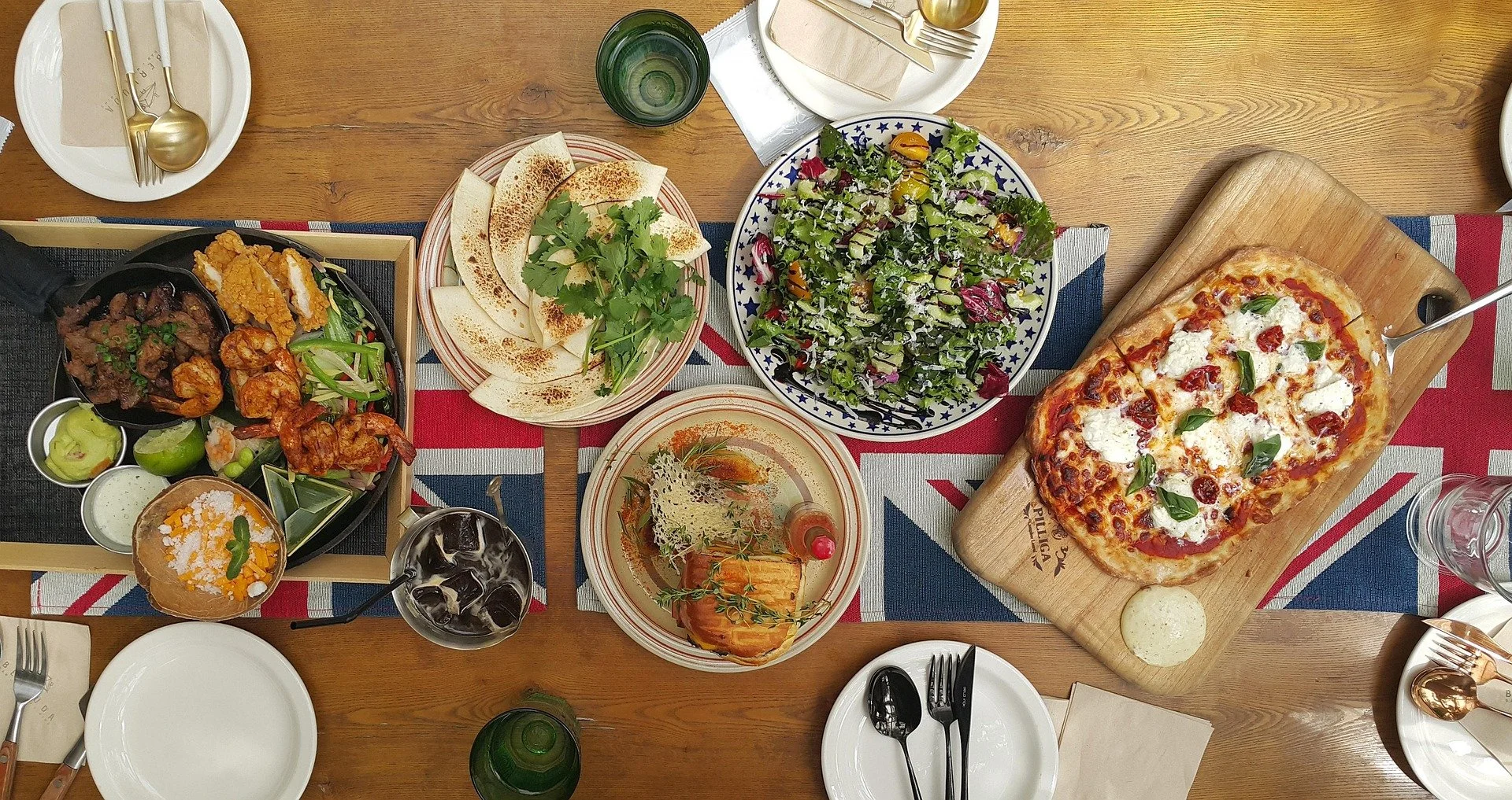A top-down view of a table set with various dishes, including a pizza on a wooden board, a salad, garlic bread, fried chicken, shrimp, and a dessert, with drinks and utensils.