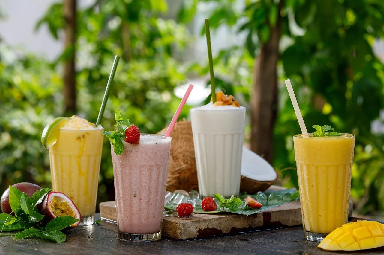 Four tropical smoothies in glasses with straws, garnished with fruits and mint, set outdoors with green trees in the background.