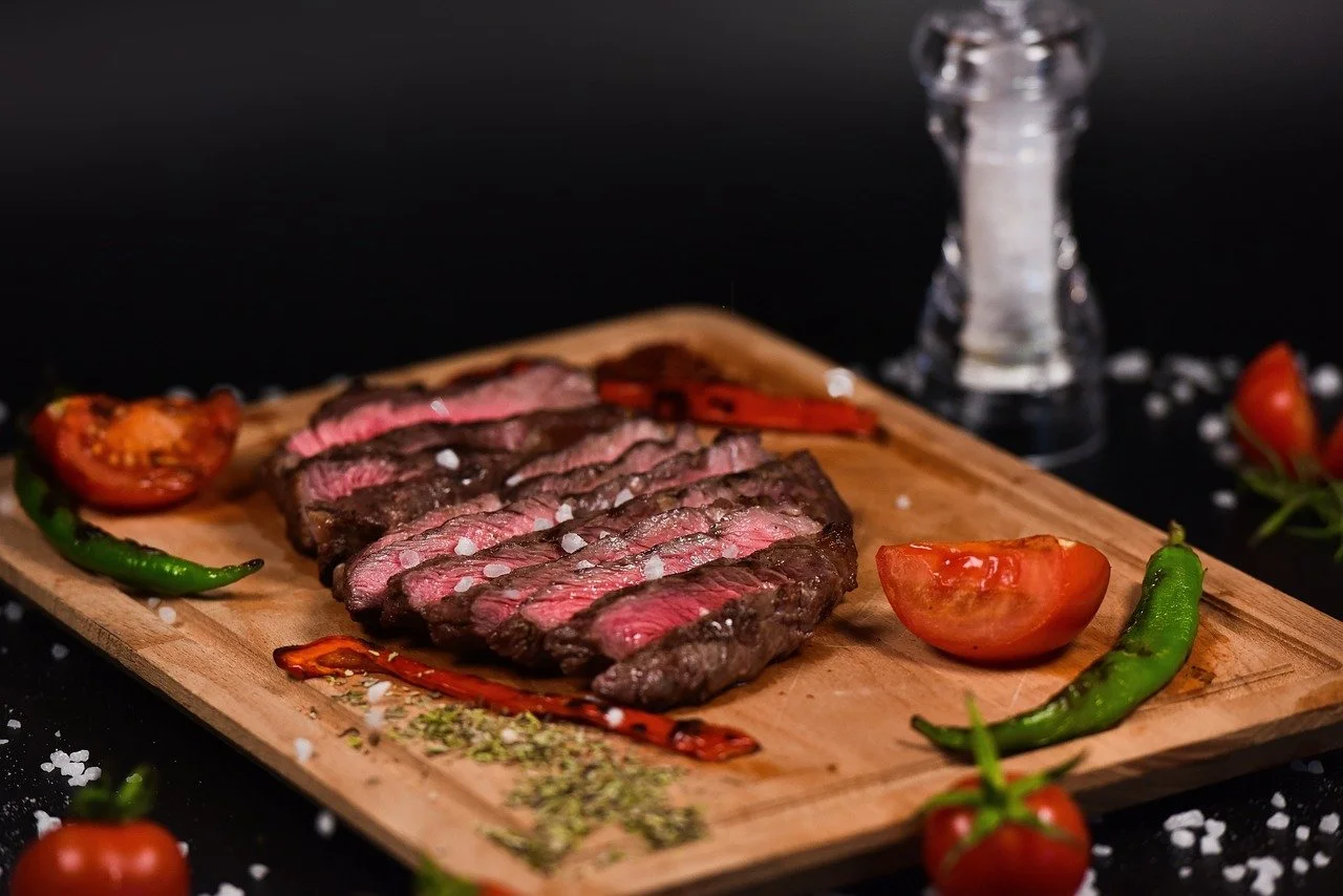 Slices of cooked steak on a wooden cutting board garnished with coarse salt. Surrounding the steak are cherry tomatoes and green chili peppers. In the background is a salt grinder, with scattered salt crystals on the black surface.