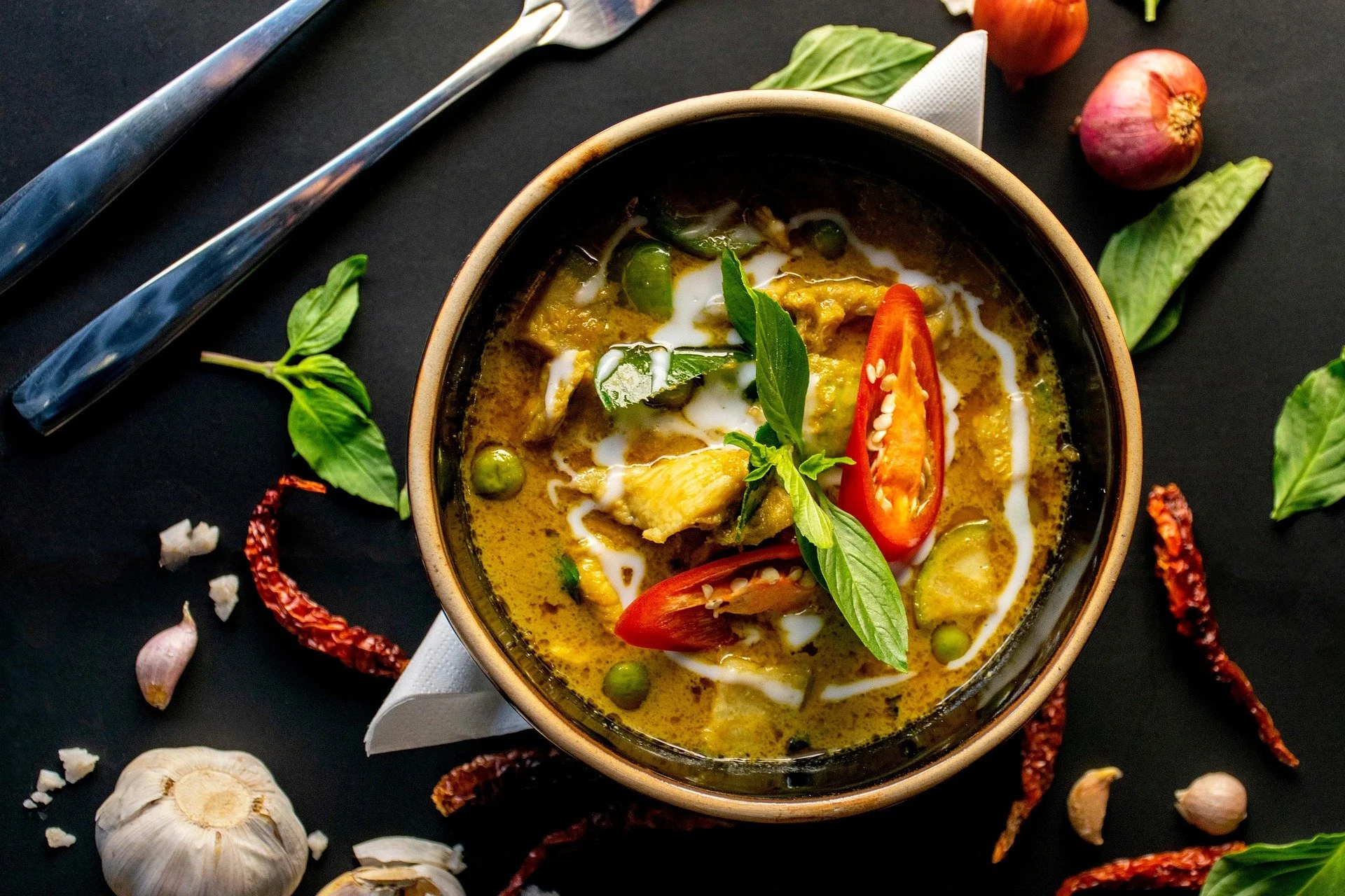 A bowl of Thai green curry with chicken, basil, red chili, and coconut milk, surrounded by garlic, dried chili peppers, and fresh herbs on a black surface.