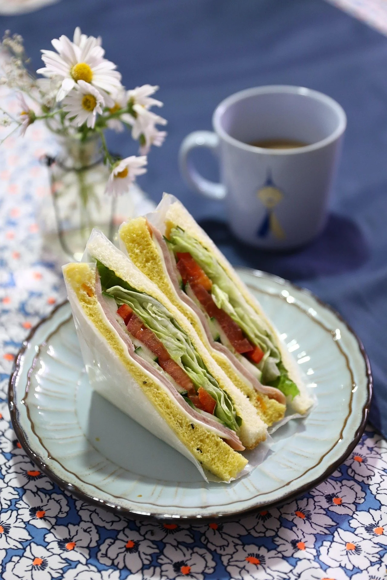 Close-up of a sandwich with lettuce, tomato, ham, and cheese on white bread, cut into two triangles, on a decorative plate. In the background, there is a cup of coffee and a small bouquet of daisies on a floral tablecloth.