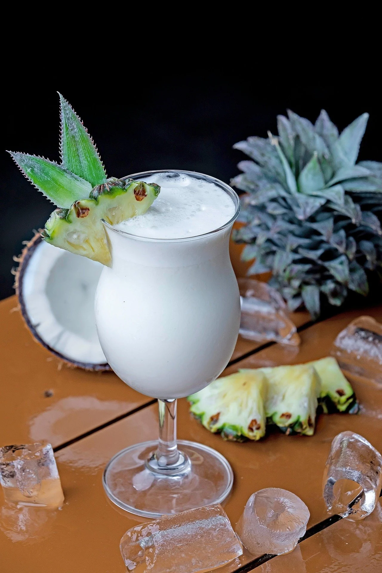 A tropical cocktail garnished with pineapple slices and a pineapple leaf, served in a tall glass with ice cubes and a halved coconut in the background, on a wooden surface with ice chips and a pineapple plant.