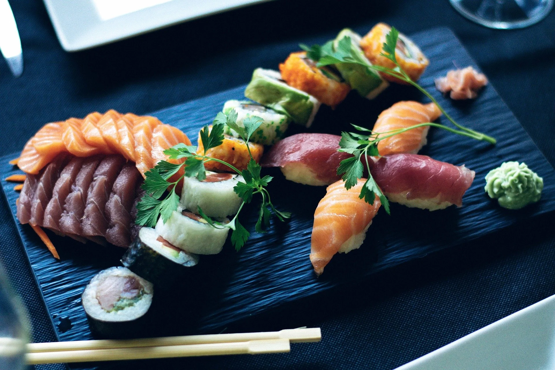 Assorted sushi platter with salmon, tuna, avocado, cucumber, and other ingredients garnished with parsley, served on a black ceramic plate with wasabi and chopsticks.