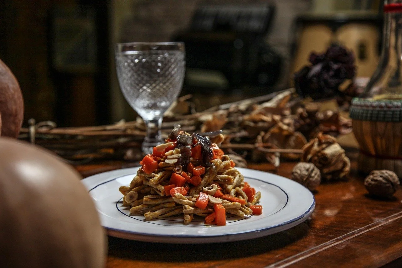 A plate of pasta with vegetables and sauce, set on a wooden table with decorative dried twigs and walnuts, in a cozy rustic setting with a glass and blurred background.