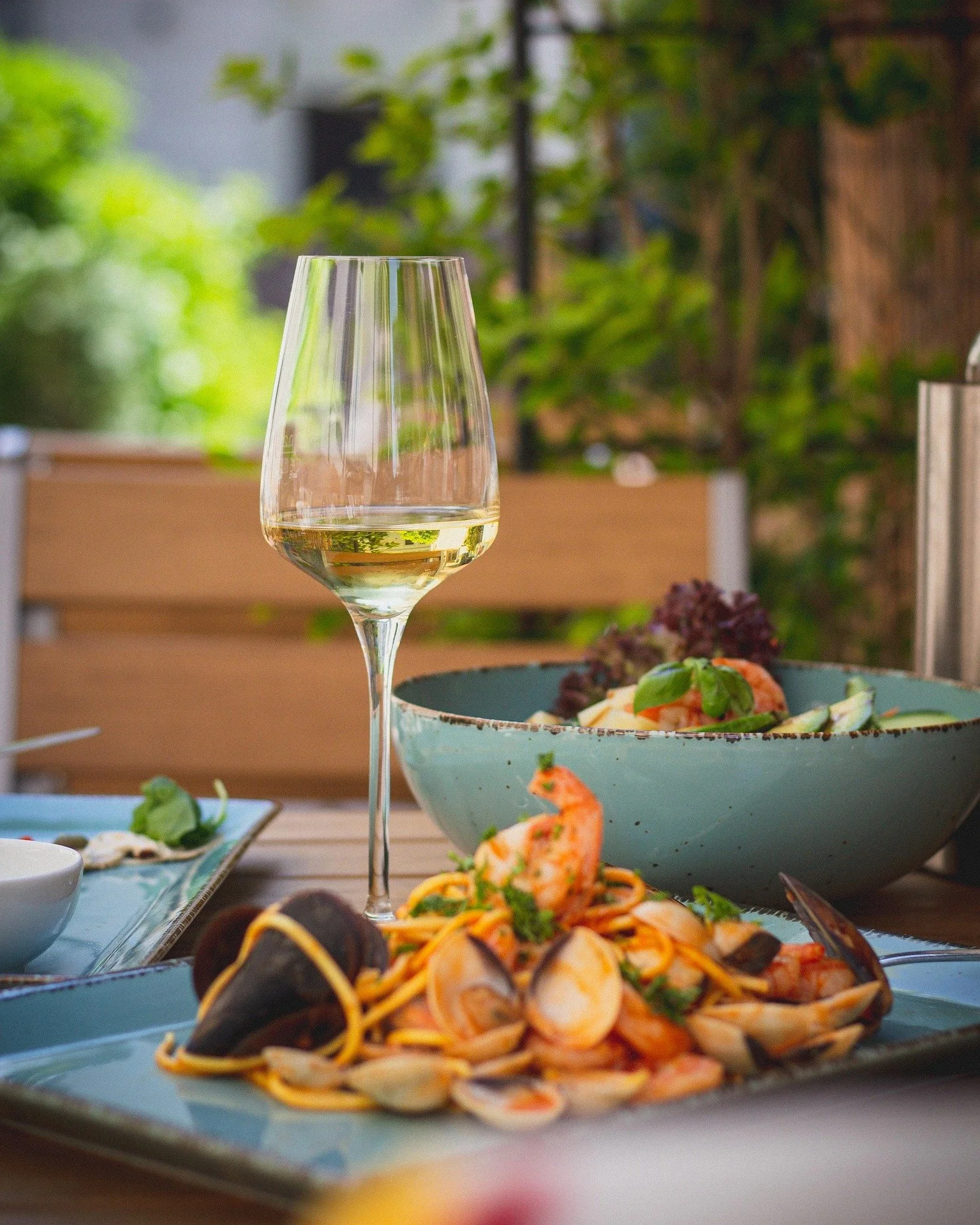 A glass of white wine and a seafood pasta dish on a table outdoors with a wooden fence and green foliage in the background.
