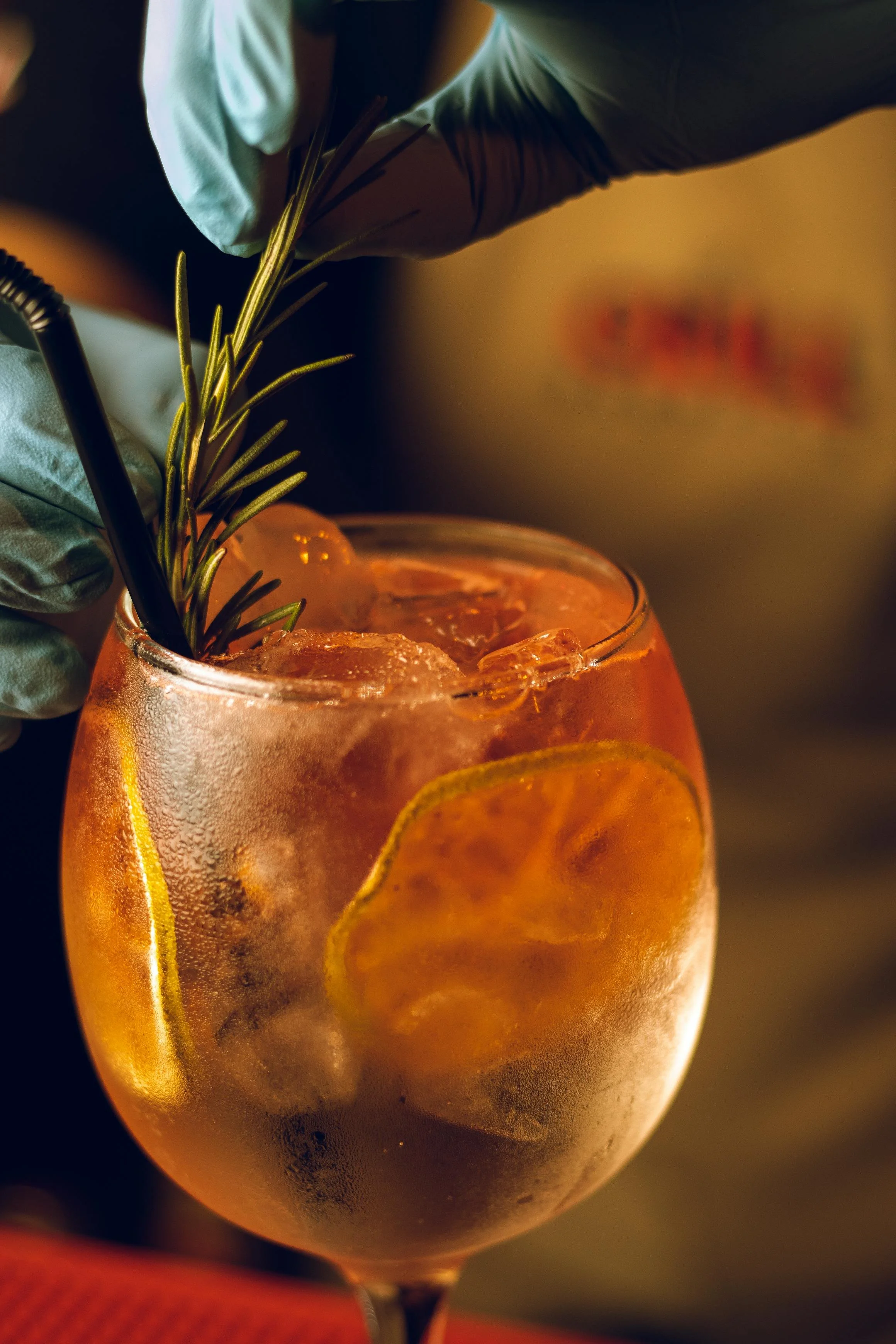 A close-up of a large glass filled with an orange-colored cocktail, ice cubes, and lemon slices, garnished with a sprig of rosemary, with someone wearing gloves adding the garnish.