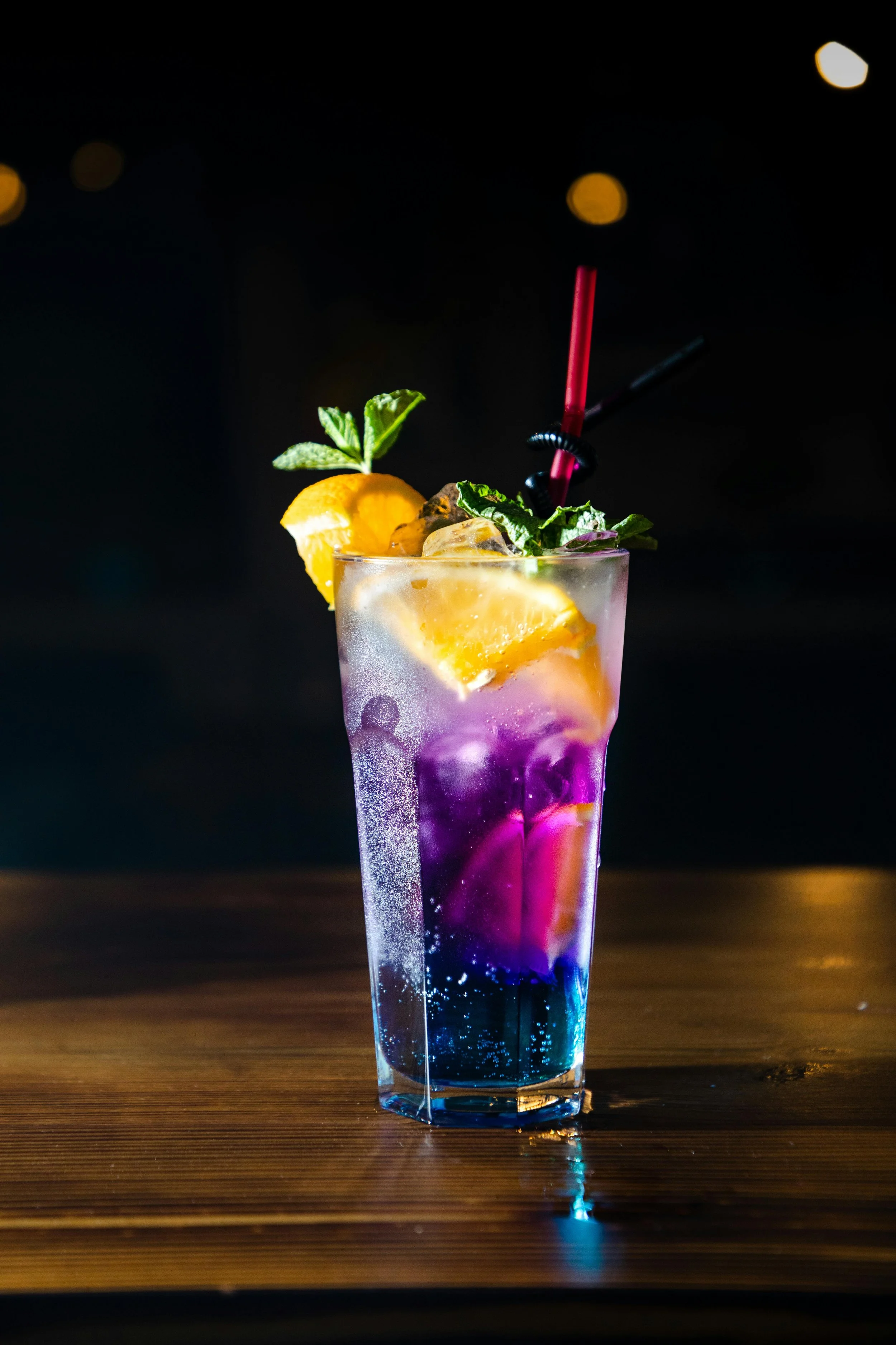 Colorful cocktail in a tall glass with lemon slices, mint leaves, and two straws on a wooden surface, against a dark background with blurred lights.