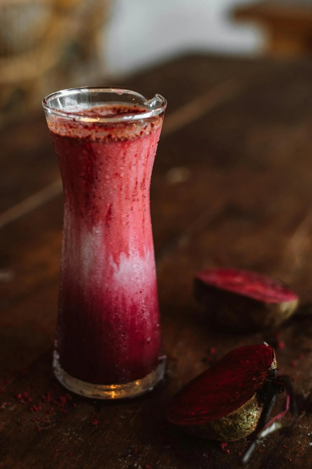 A tall glass of smoothie with a gradient from deep red at the bottom to a lighter pink at the top, placed on a dark wooden surface next to halved cacao pods.
