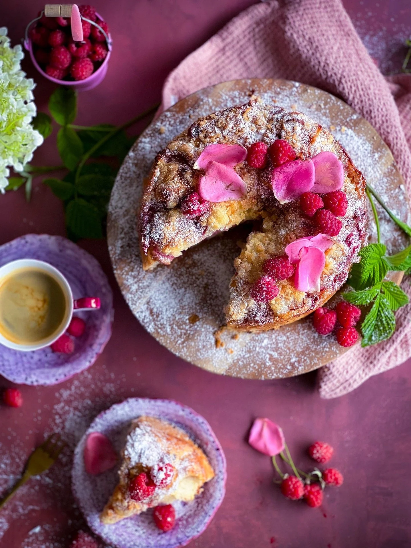 A whole Bundt cake topped with powdered sugar, raspberries, and pink rose petals, with one slice cut out. The slice and some raspberries are on separate plates, and a cup of coffee is nearby. Fresh flowers and berries are also present on the table.