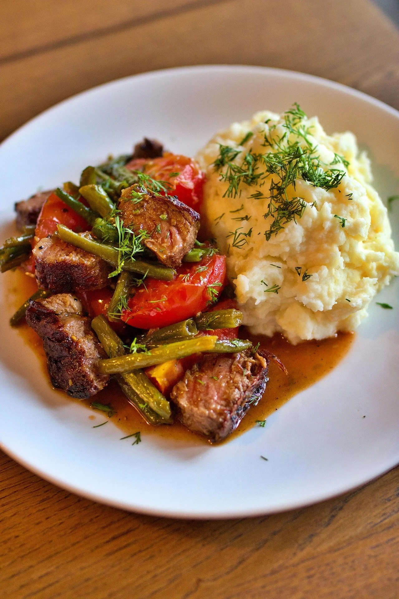 A white plate with mashed potatoes garnished with chopped herbs on the right, and a beef and vegetable stew with gravy on the left, served on a wooden table.