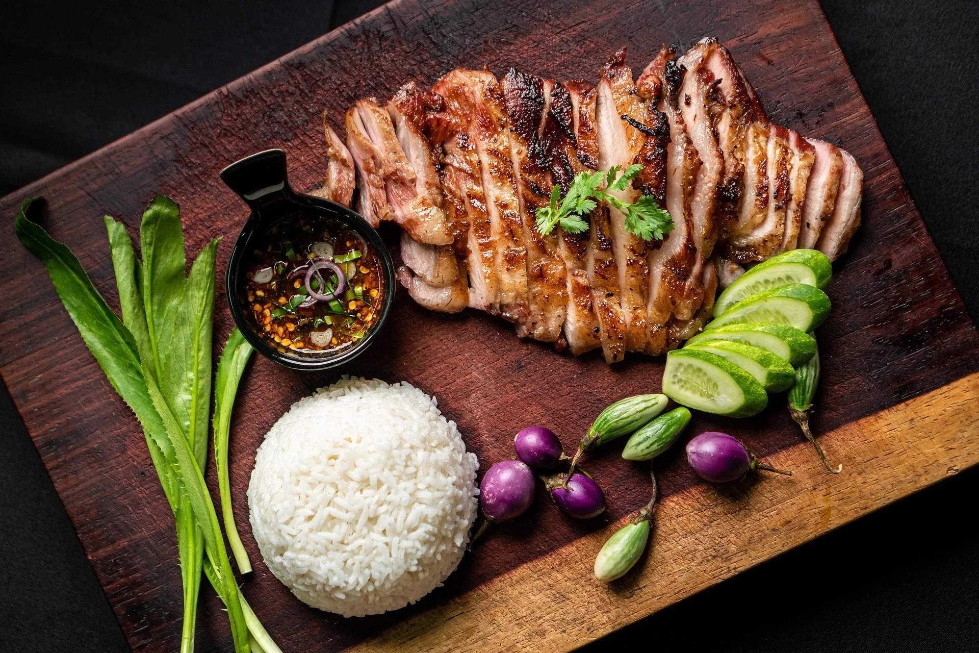 Grilled sliced pork belly with fresh herbs, cucumber slices, purple eggplants, steamed white rice, and dipping sauce on a wooden platter.