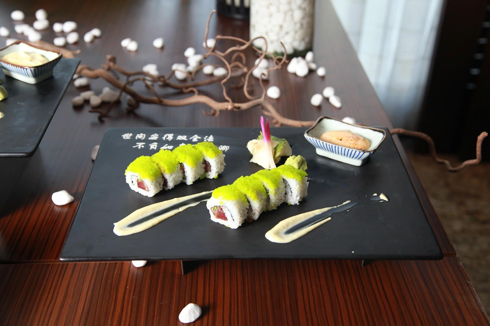 Sushi rolls with green topping on a black rectangular plate, with cream sauce drizzled around, accompanied by dipping sauce in a small bowl, on a dark wooden table decorated with white shells and twisted branches.