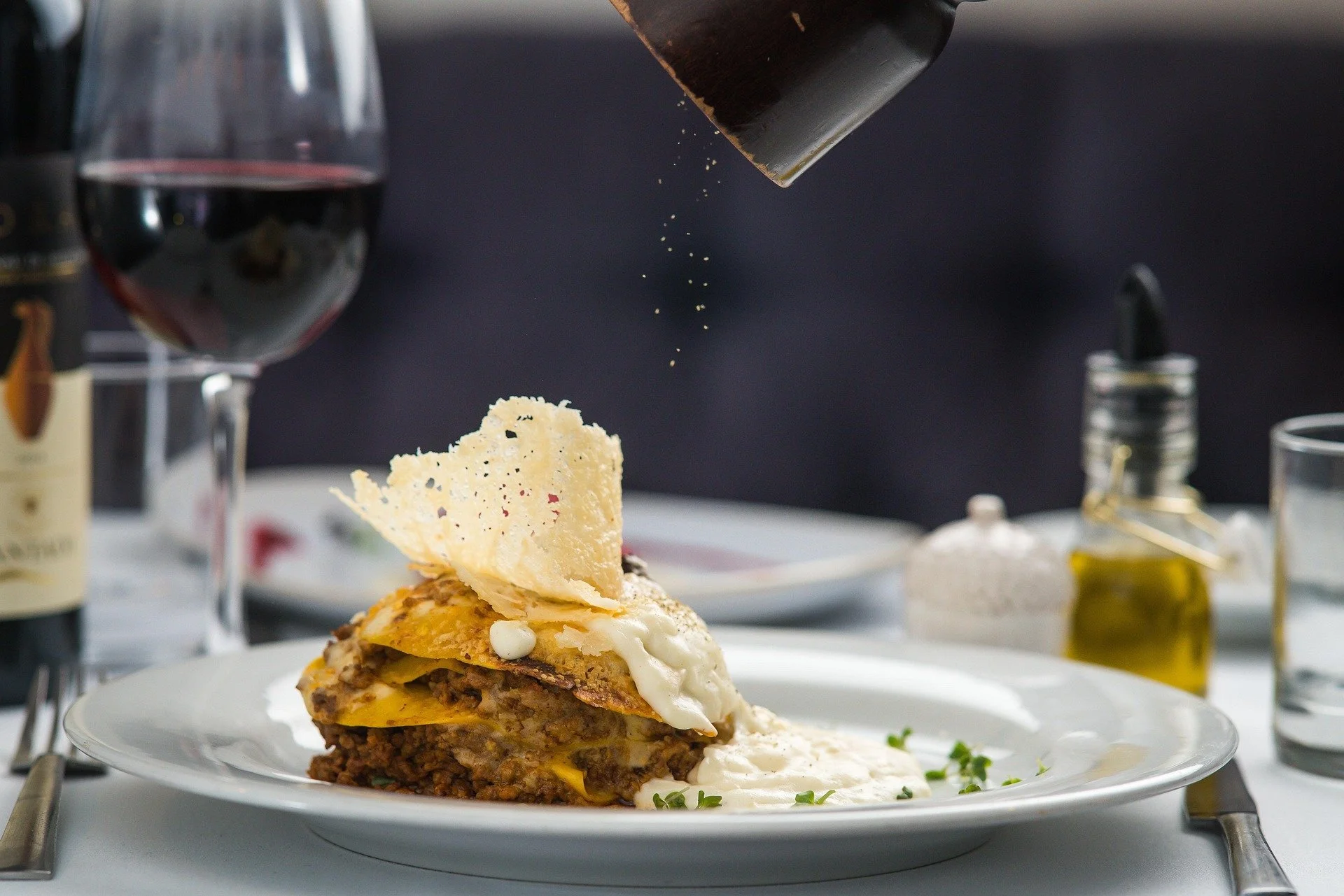 A plate of layered lasagna topped with cheese and a Parmesan crisp, with salt and pepper shakers, a glass of red wine, olive oil, and salt on the table.