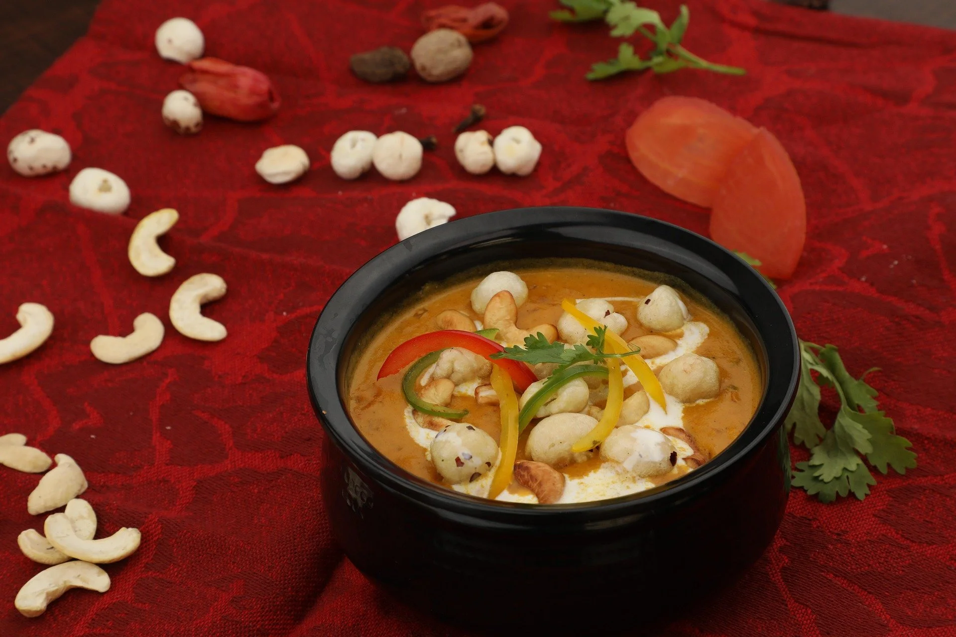 A bowl of Indian curry garnished with green coriander, served with slices of tomato and yellow and green bell peppers, placed on a red cloth with scattered cashew nuts, dried fruits, and cilantro leaves around it.