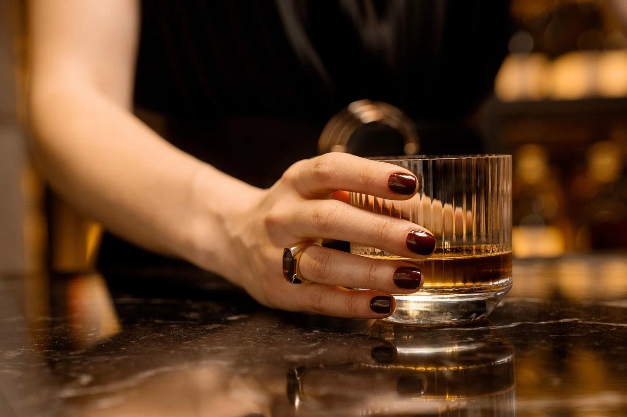 A woman with dark nail polish holding a glass of whiskey on a dark marble bar counter.