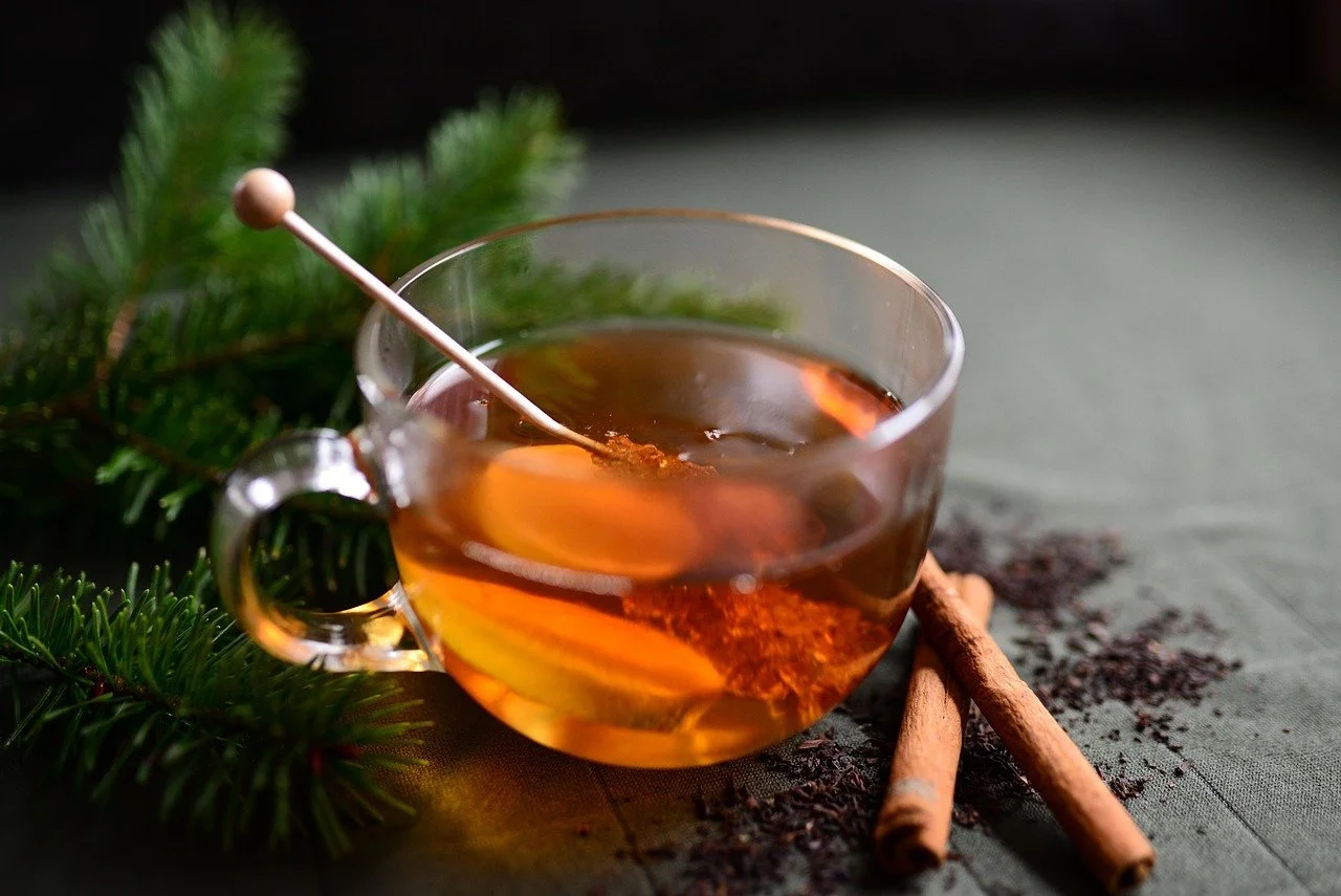 A glass cup of hot tea with a cinnamon stick on a table, accompanied by some loose cinnamon and a sprig of pine branches for decoration.