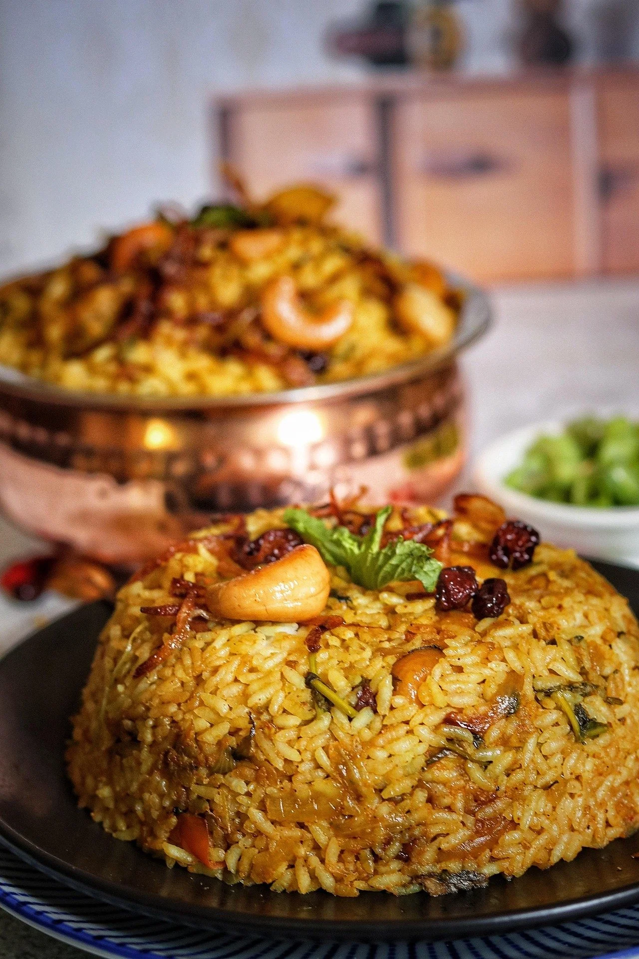 A plate of fried rice garnished with cashews, dried cranberries, and cilantro, with a bowl of salad in the background.