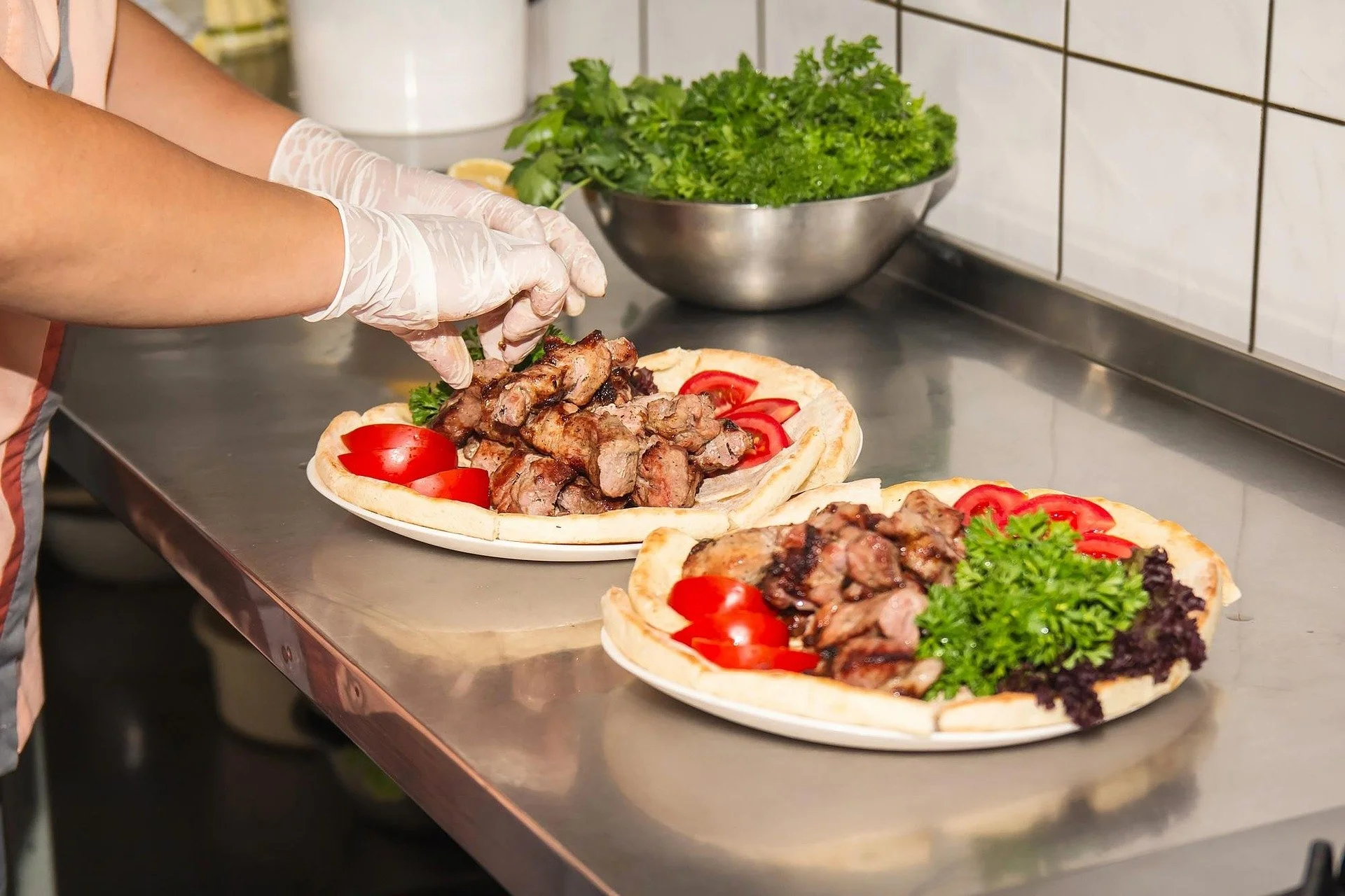 A person wearing plastic gloves preparing a flatbread pizza topped with grilled meat, cherry tomatoes, and greens on a stainless steel countertop, with a bowl of fresh herbs in the background.
