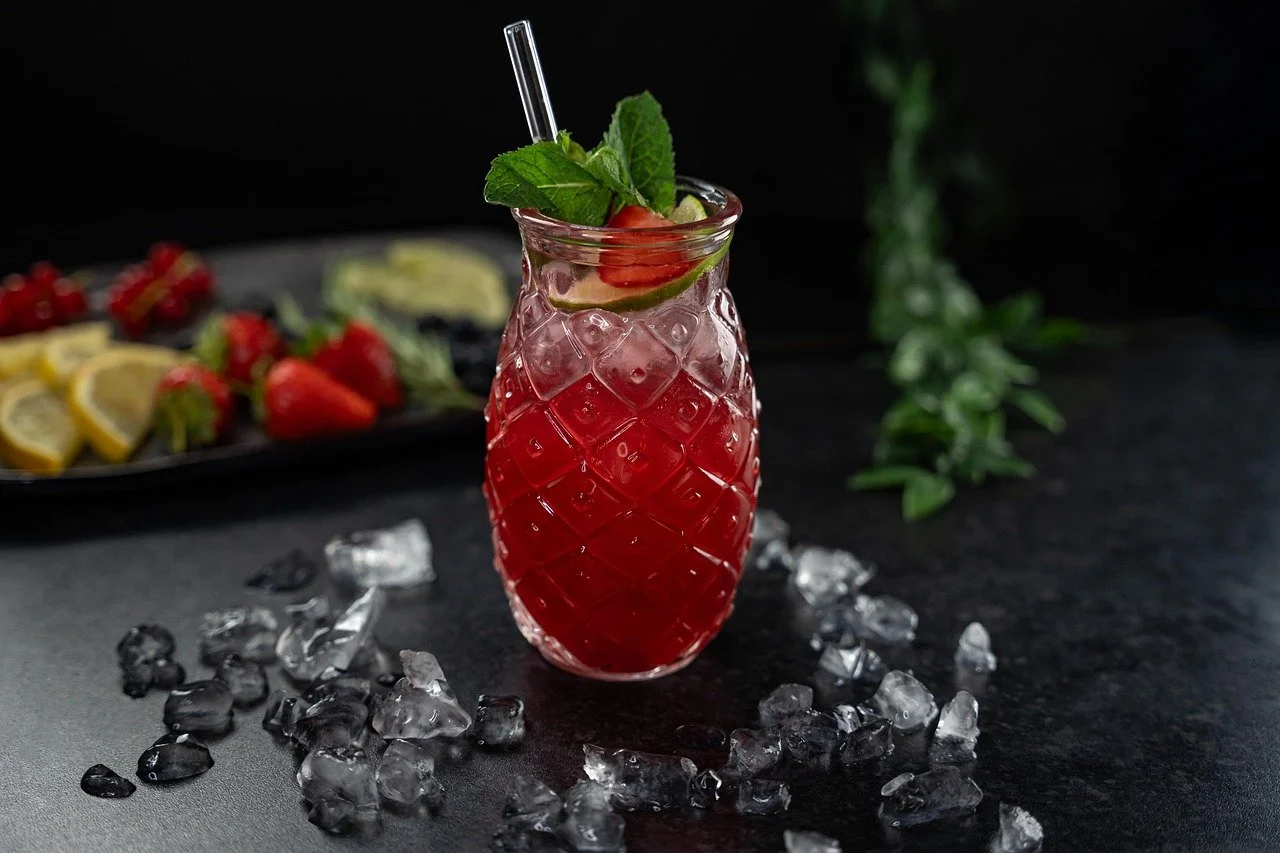 A glass tiki mug filled with a red cocktail, garnished with a mint sprig, a lime slice, and a strawberry, surrounded by ice cubes on a dark surface. In the background, a platter of lemon wedges, strawberries, and blueberries.
