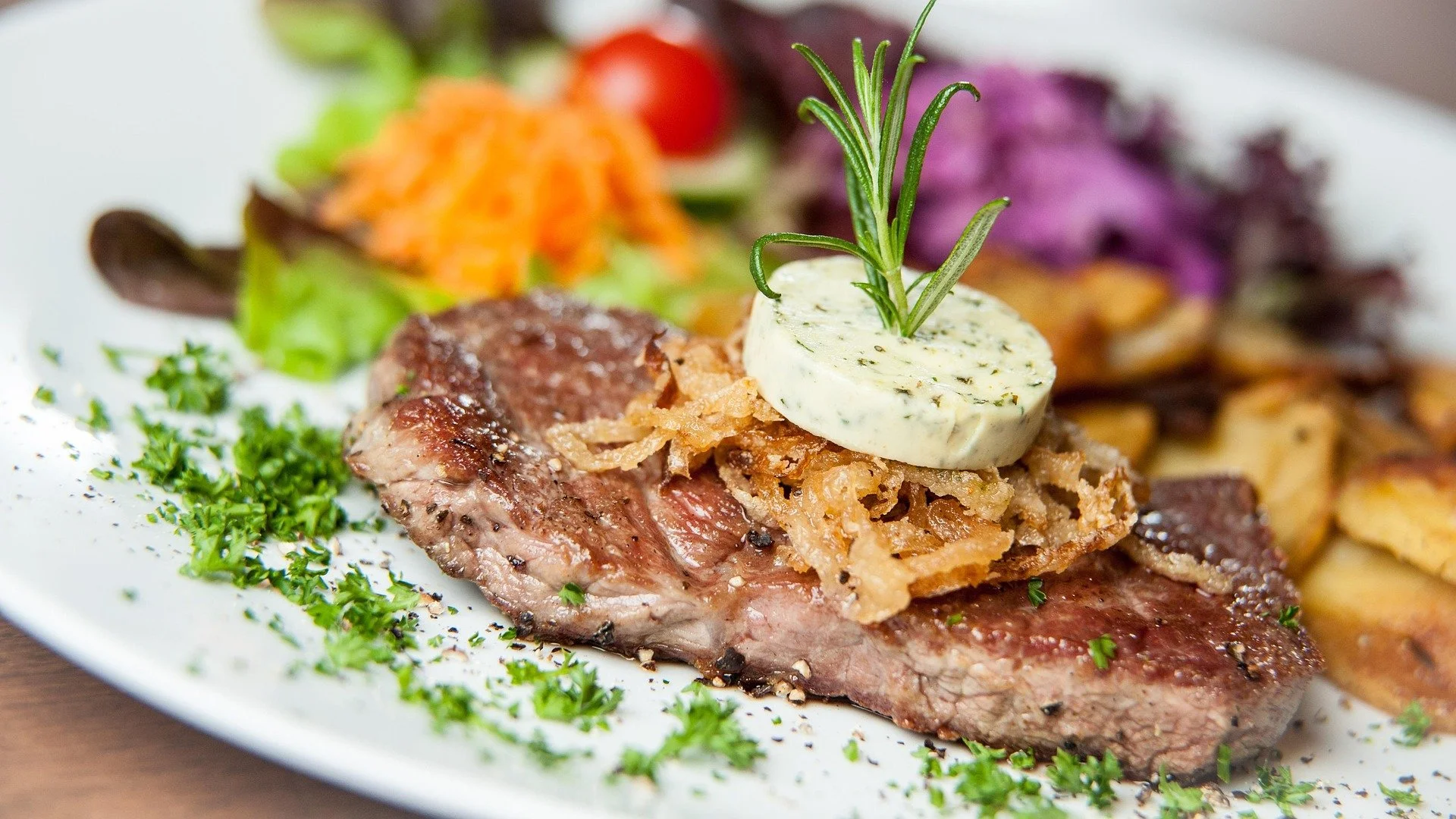 Close-up of a plated meal with grilled steak topped with onion straws, a dollop of herb butter garnished with a sprig of rosemary, and side dishes including shredded carrots, cherry tomatoes, purple cabbage slaw, and diced fried potatoes.