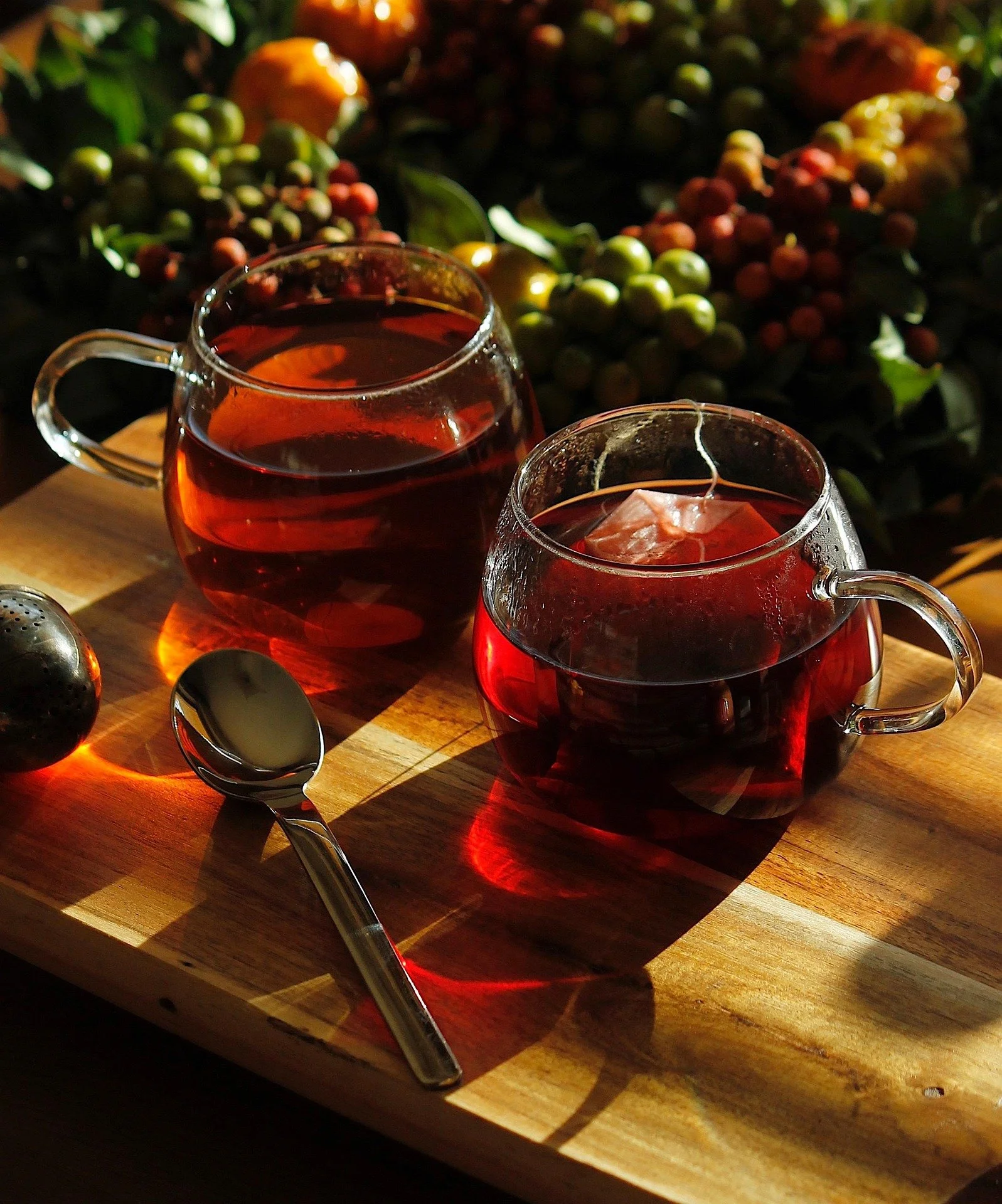 Two glass cups of brewed tea on a wooden cutting board, with a silver spoon and a black fruit near them, and a background of green leaves and colorful berries.
