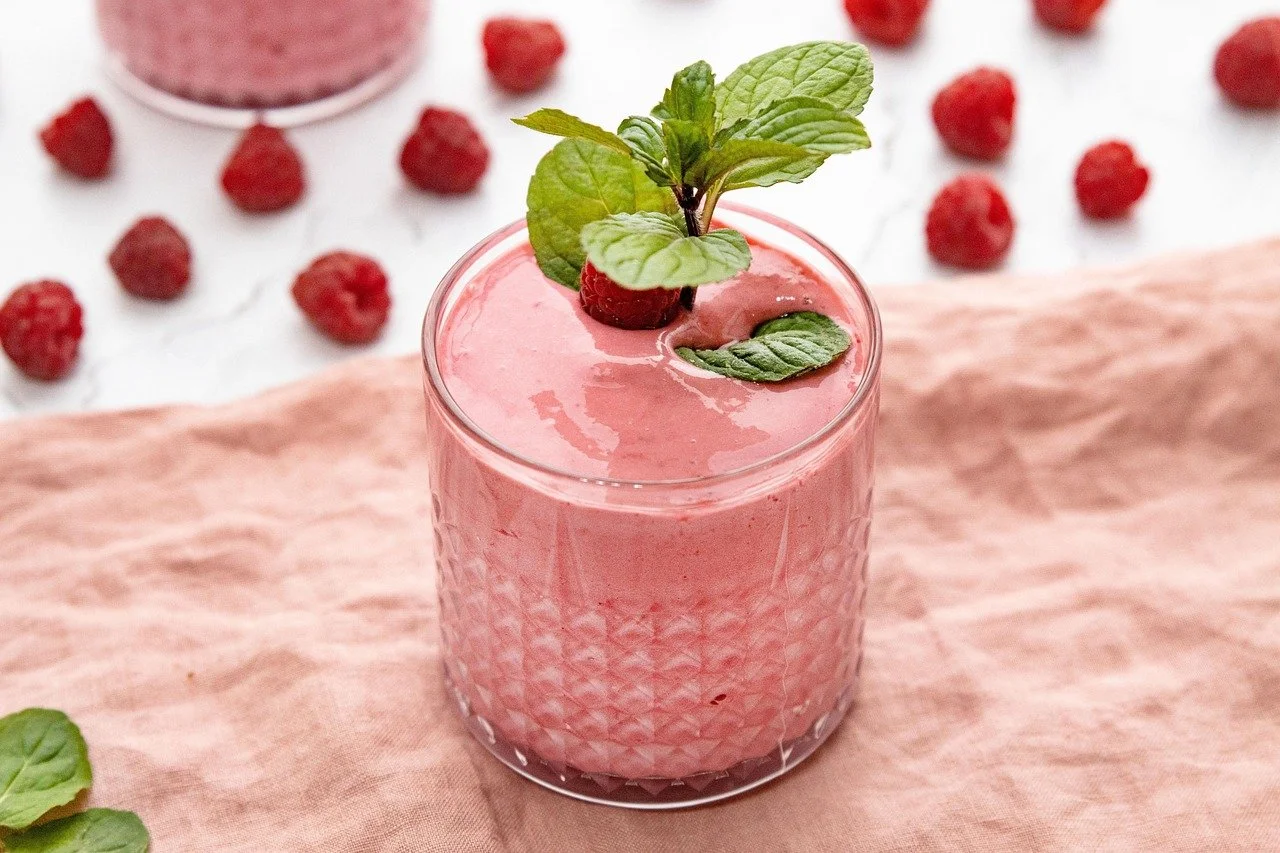 Pink raspberry smoothie garnished with fresh mint leaves, in a glass on a pink cloth, with raspberries scattered in the background.