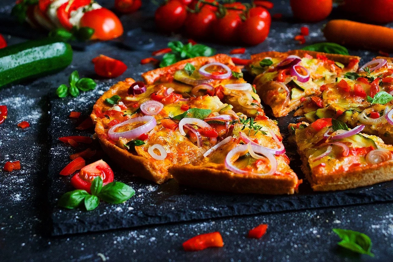 A freshly baked vegetable pizza sliced on a black surface, topped with onions, tomatoes, bell peppers, and herbs, with cherry tomatoes, a cucumber, and fresh basil scattered around.