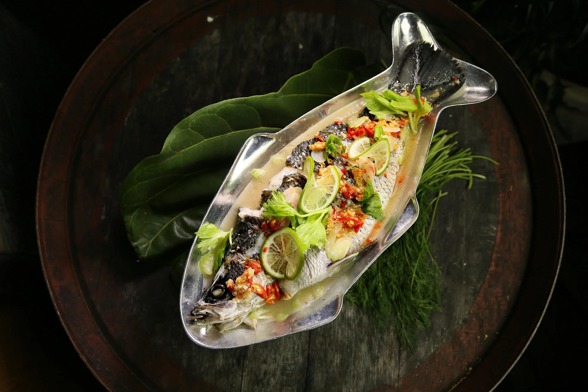 Whole fish garnished with lime slices, herbs, and chopped vegetables on a silver platter, placed on a wooden surface with green leaves.