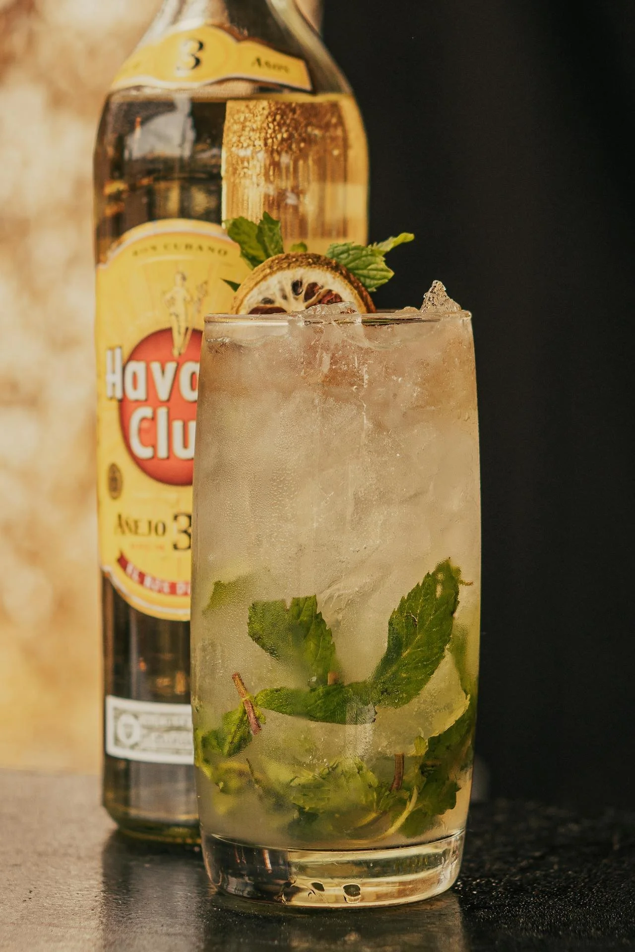 A tall glass mojito cocktail garnished with mint leaves and a dried lime slice, with a bottle of Havoc Club Añejo 3 Años rum in the background.