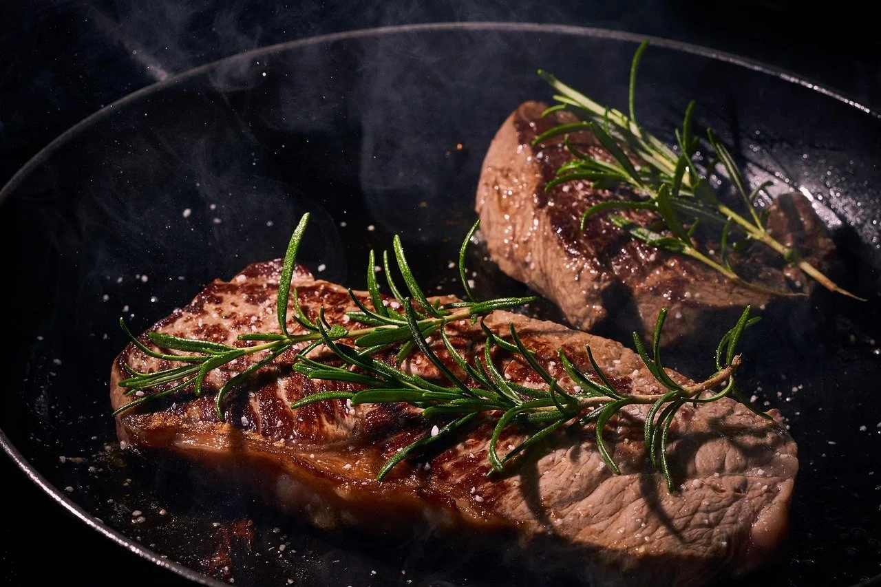 Two seasoned steaks cooking in a black skillet with sprigs of fresh rosemary on top.