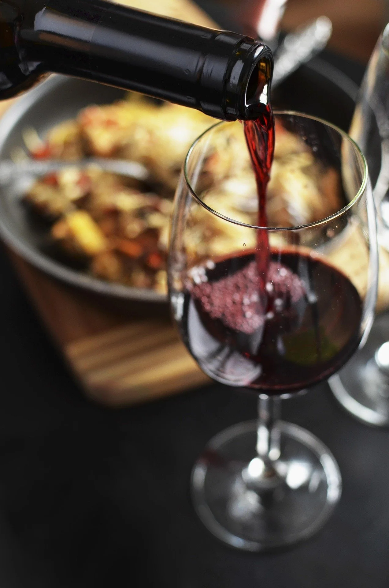 A glass of red wine being poured from a bottle, with a dish of food in the background.