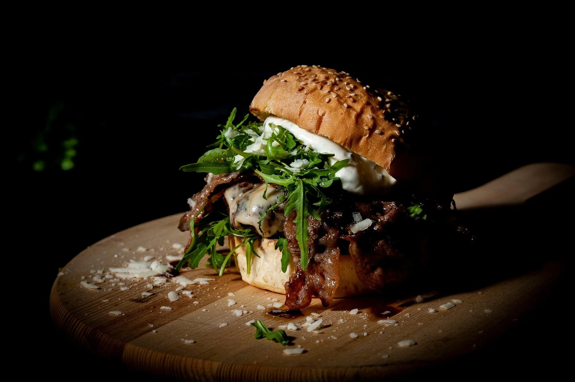 Close-up of a gourmet burger with bacon, arugula, cheese, mayonnaise, and a sesame seed bun on a wooden serving board against a dark background.
